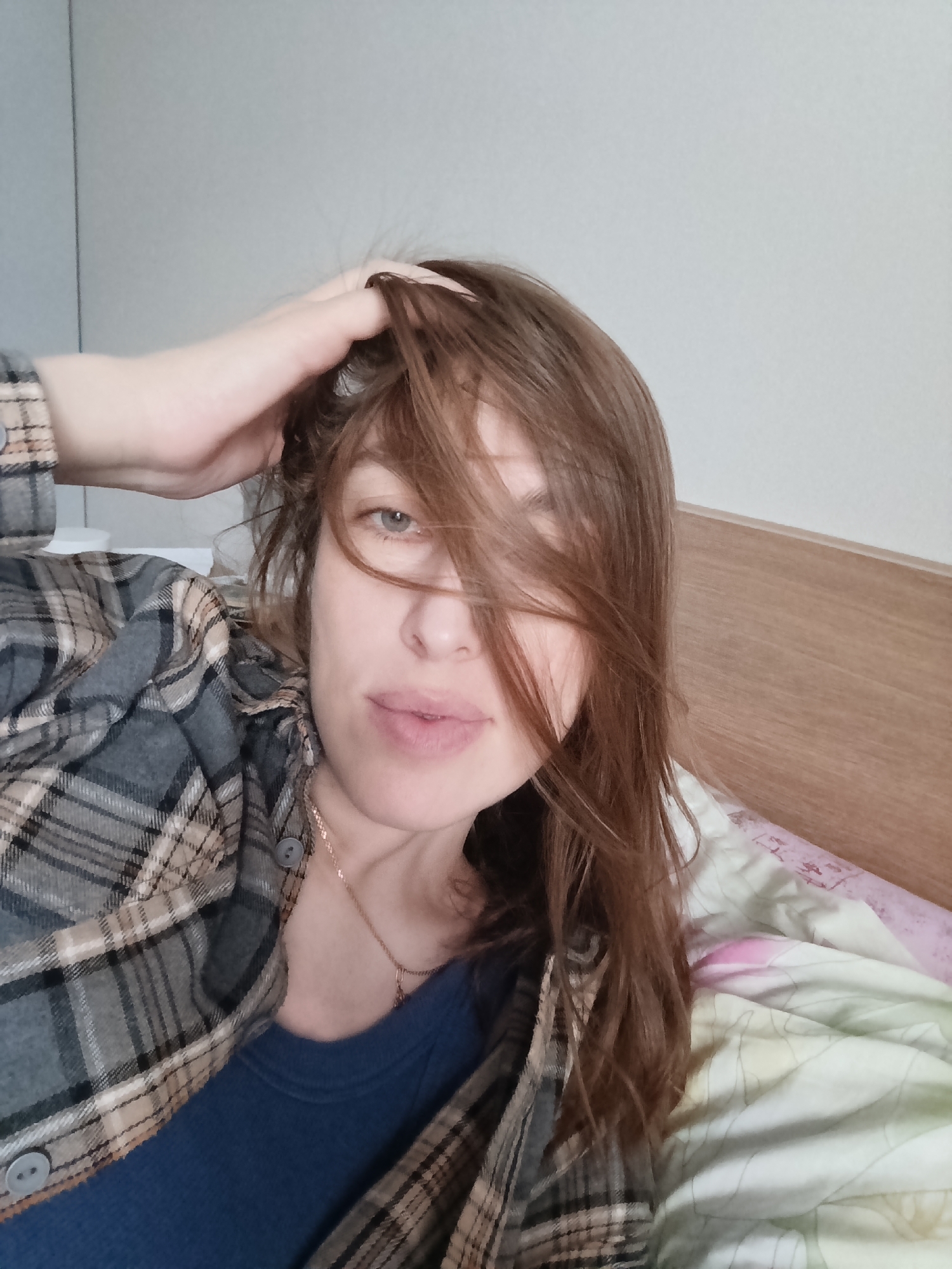 A woman lies on a bed with a wooden headboard, a floral-patterned blanket, and a light-colored wall. She wears a blue shirt under a gray and tan plaid flannel. Her left hand is on her head, lifting her brown hair, partially obscuring her face. A gold necklace is visible around her neck. The location is Chaikovsky, Russia.
