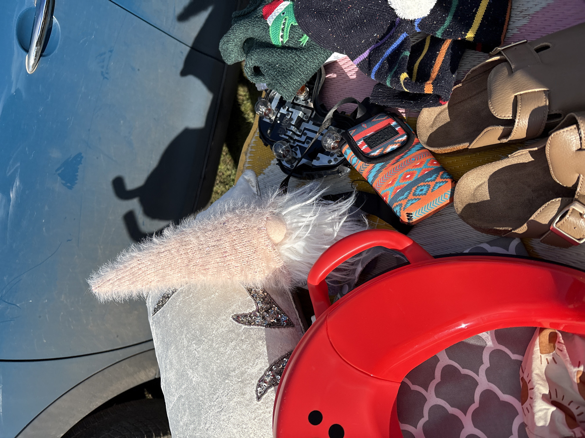 The image shows various objects placed on a yellow striped mat next to the blue car. There's a light pink gnome, some hats, a pair of brown baby shoes, a black taillight and a red toy.
The location is not specified, though the prompt stated it was in the city of Chirk, United Kingdom.