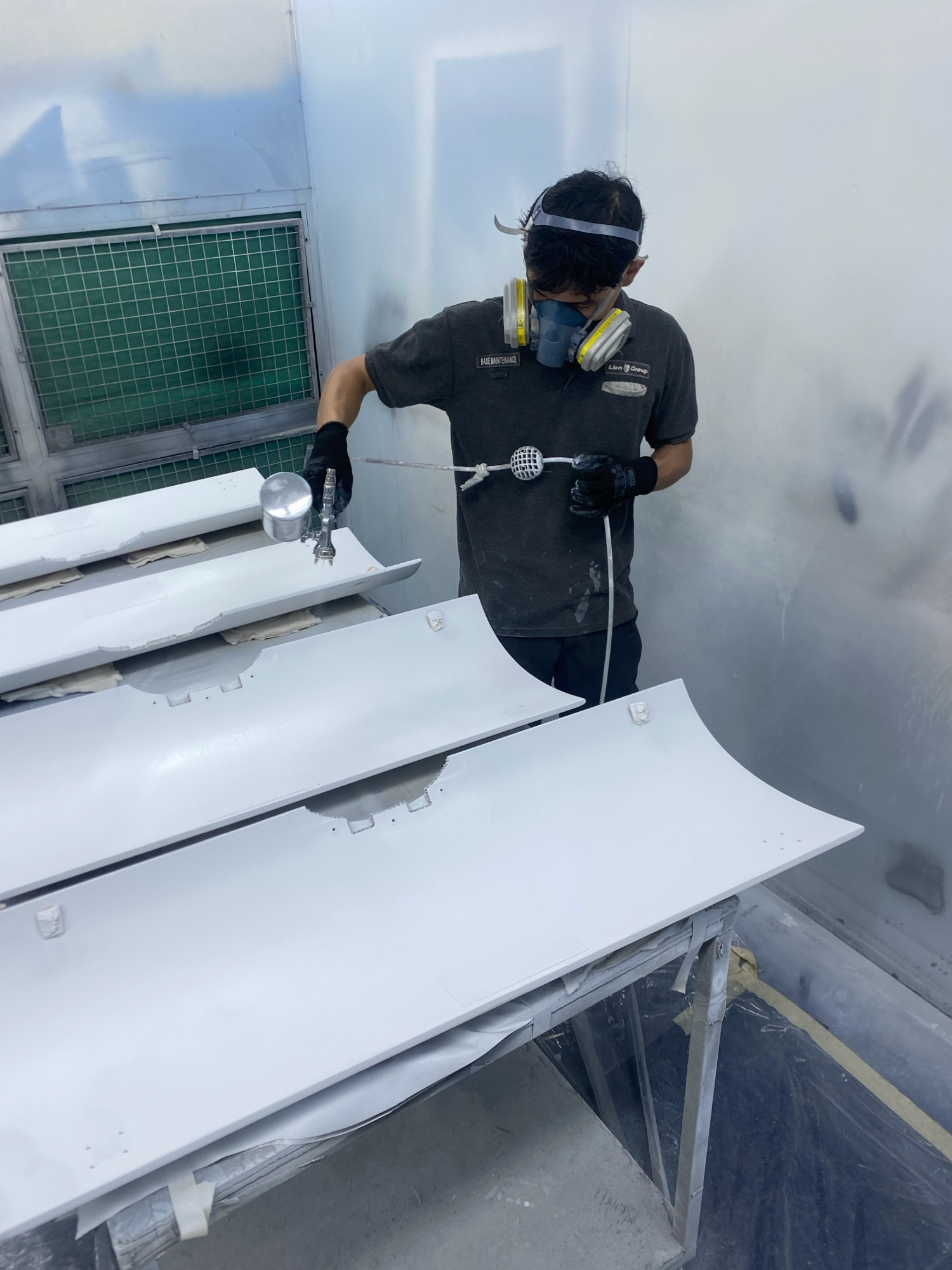 A person wearing a respirator mask and gloves is spray-painting white panels in a paint booth. The person is holding a spray gun and aiming it at the panels, which are laid out on a table. The paint booth is a clean, well-lit space with ventilation systems visible on the wall. The panels appear to be automotive parts, possibly car doors or body panels. The scene is a workshop or factory environment in Batam, Indonesia.