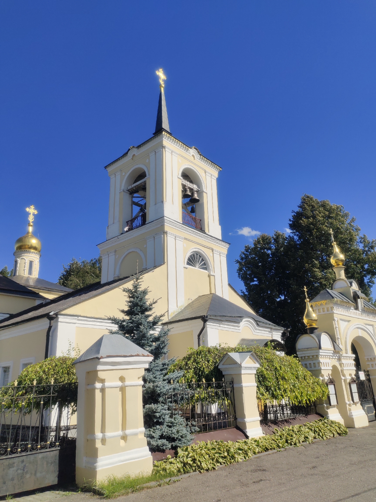 This is a low-angle outdoor photograph of a yellow church with a prominent bell tower and golden domes against a clear blue sky. The church features arched windows and decorative moldings. In the foreground, there is a black metal fence and a light yellow pillar with decorative elements. Green bushes and trees are visible behind the fence and surrounding the church. The sun is shining, casting shadows and highlighting the golden crosses on the domes. The image appears to be taken in Ostashkovo, Russia, given the architectural style suggestive of Russian Orthodox churches. There are no people visible in the frame, and no actions or interactions are taking place. The environment is peaceful and sunny.