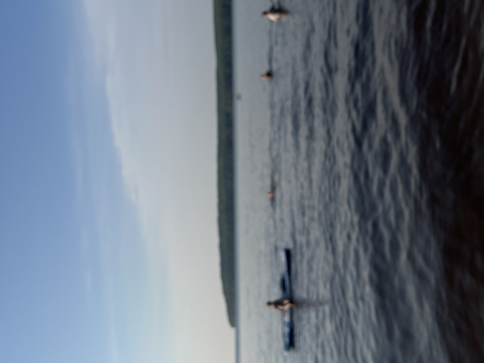 A group of people are enjoying recreational activities on the Kama River in Chaikovsky, Russia. The scene depicts a person sitting on a paddleboard in the foreground, facing towards the left. Further in the distance, several other individuals are visible in the water, either swimming or wading. The background features a tree-lined shore, suggesting a natural landscape surrounding the river. The sky is clear with some light clouds, indicating fair weather. The overall impression is one of leisure and outdoor enjoyment.