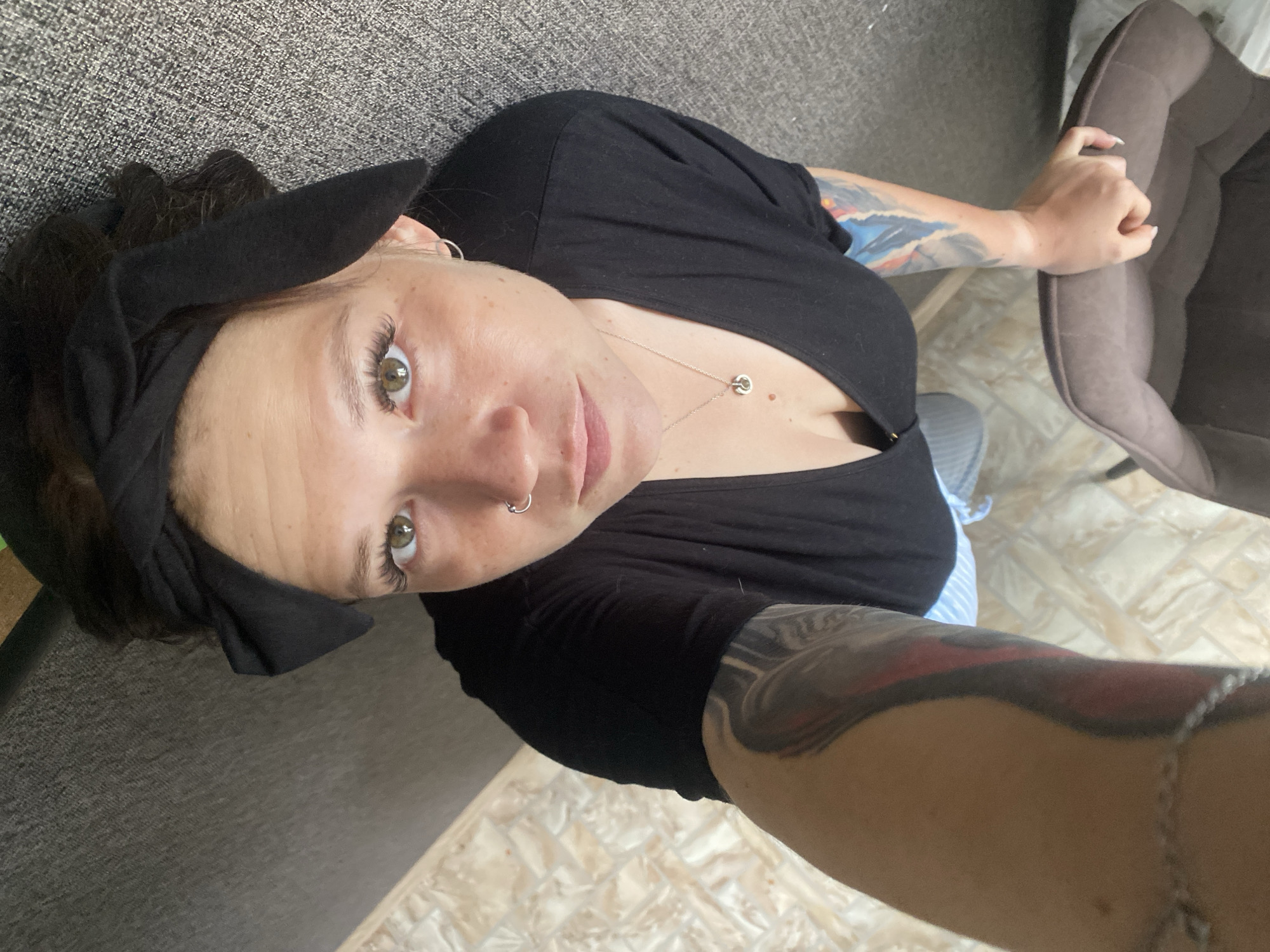 A high-angle, full shot shows a woman from the chest up, wearing a black v-neck shirt, a black headband, and light-colored striped pants. She has green eyes, a nose ring, and a necklace with a small circular pendant. She has extensive tattoos on her left arm and a tattoo on her right arm. Her left hand is visible at the bottom of the frame, resting on a brown object that appears to be a dog bed. The background is a textured gray surface, possibly upholstery or carpet, and a tiled floor. The lighting is bright and appears to be natural.