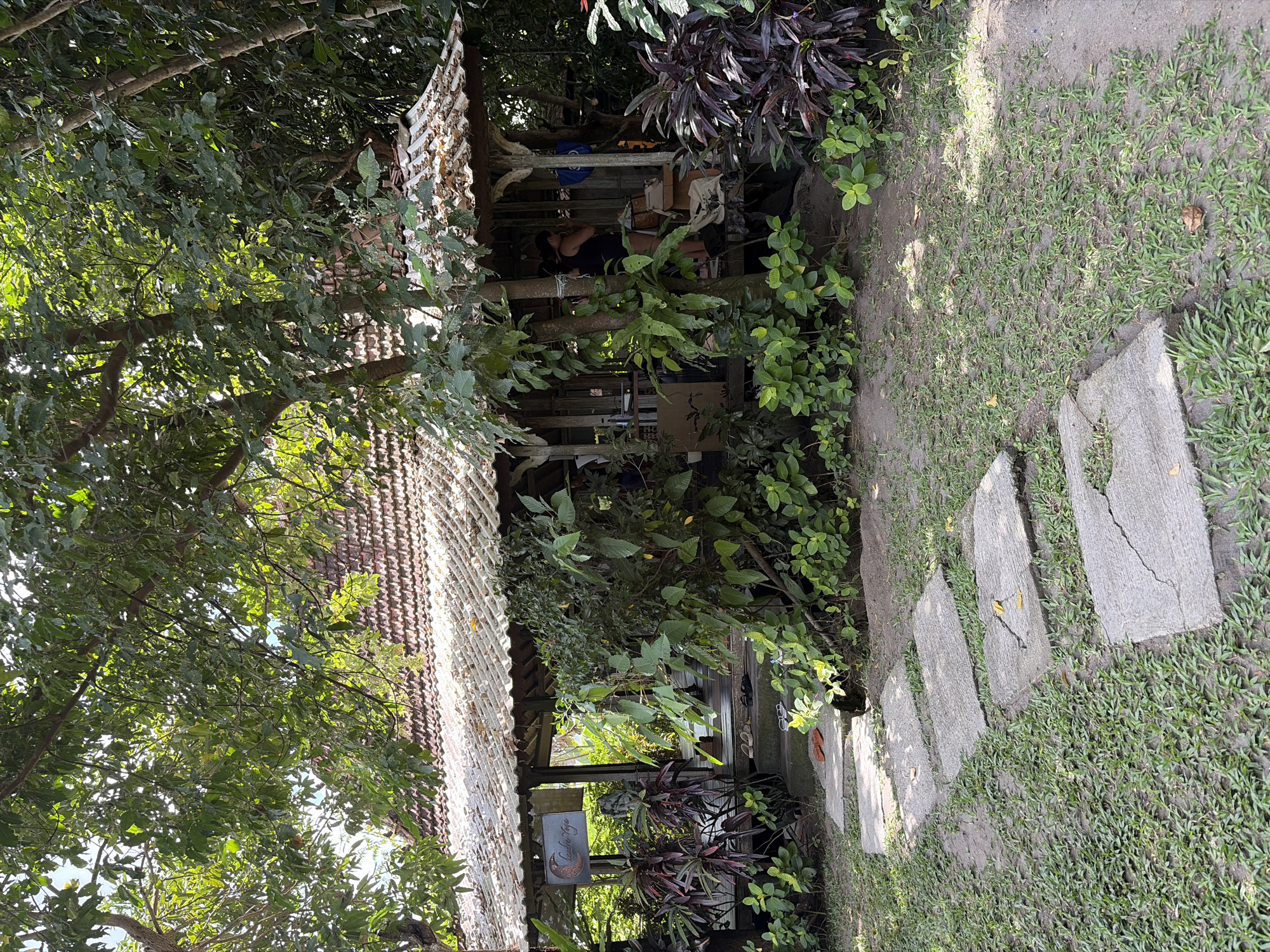 The image depicts an outdoor scene at a location in Mengwi, Indonesia, featuring a small, open-air structure with a tiled roof, possibly a cafe or a shop. The structure is surrounded by lush greenery, including trees and various plants with dark red and green foliage. A stone pathway with several rectangular flagstones leads from the foreground towards the structure. The pathway is laid on a patch of ground covered with short grass and some sand. The structure itself appears to be made of wood and has an open front, revealing a counter area inside. Two people are visible within the structure, one wearing a black tank top and the other partially obscured. There are several pairs of flip-flops placed on the steps leading up to the structure. A sign with a crescent moon and text is hanging on the left side of the entrance. The overall environment suggests a tropical setting, likely a tourist-oriented establishment.