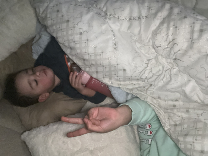 A child sleeps soundly on a pillow, clutching a pink water bottle, partially covered by a patterned white comforter. Another person’s hand emerges from under the comforter, displaying a "peace" sign. Their arm, clad in a mint green Champion sweatshirt, is also visible. The sleeping child is resting on a tan sofa.