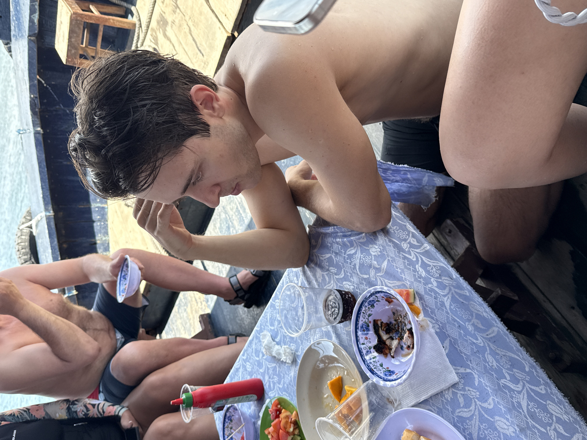 On a boat in Nha Trang Ward, Vietnam, several partially visible men sit around a table covered with a patterned blue cloth. One man with dark, wet hair leans over the table, his arms resting on it. Plates of food and empty cups are scattered across the table. The men wear swim attire. The boat is near the water.