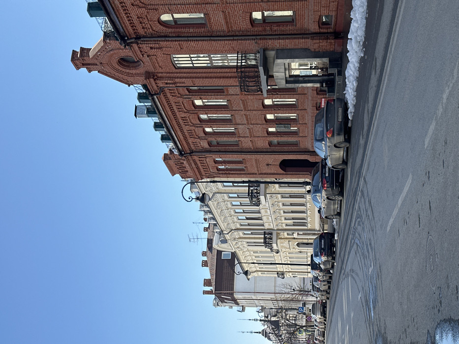 The image displays a street scene in Kazan, Russia, under a clear blue sky. A row of multi-story buildings lines the right side of the paved thoroughfare, extending into the distance.
In the immediate foreground on the right, a prominent red brick building with ornate brickwork, arched windows, and a distinctive gable is visible, featuring an elevated entrance beneath an iron balcony. Adjacent to this, a dark-colored sedan (license plate X 781 XX 116) and a darker sedan (license plate K 010 HC 116) are parked curbside, with patches of snow and slush between the vehicles and the building. Further down the street, a continuous line of various passenger vehicles is parked along the curb.
To the left of the red brick structures, several cream-colored buildings with highly ornamented facades, including classical architectural details such as pilasters and decorative window surrounds, are visible. The asphalt street surface shows signs of moisture and tire tracks, with remnants of snow present along the curb and in the gutter. Two ornate black streetlights with globe fixtures are situated along the sidewalk. No individuals are visible within the scene.