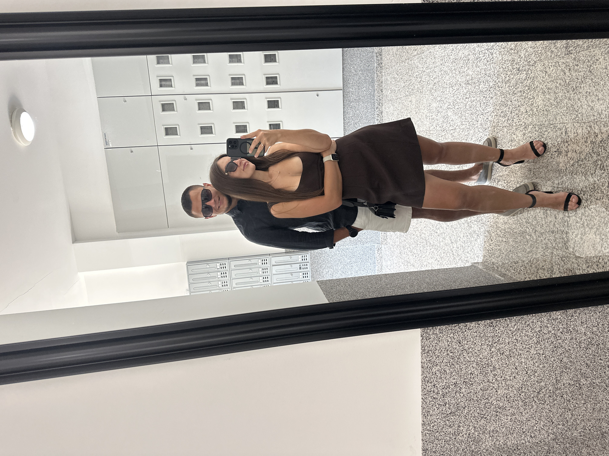 A young couple takes a mirror selfie in a modern, minimalist hallway with lockers against a white wall. The man, wearing sunglasses and a dark shirt with white shorts, stands behind a woman in sunglasses and a dark brown dress, who is holding a smartphone to capture the photo. She is wearing black heeled sandals, and he is wearing grey slip-on shoes. The floor is tiled with a speckled grey and white pattern, and the walls are white with a granite-like texture on the lower half. The lockers appear to be part of a mail or storage system. The overall impression is one of a clean, contemporary urban environment.