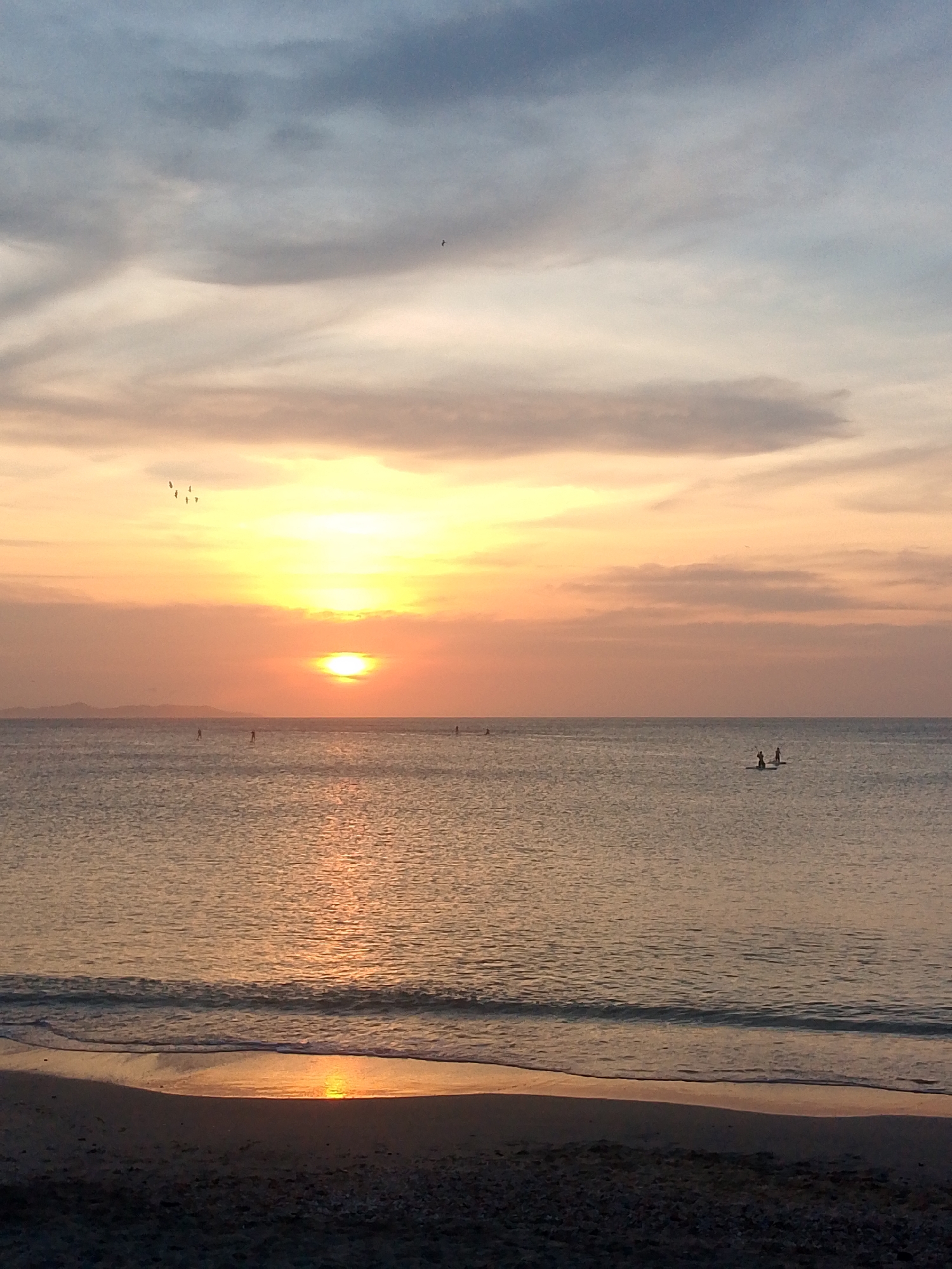 Sunset over the water in Juan Griego, Venezuela. A sandy beach stretches across the foreground, transitioning into gentle waves. In the middle distance, the sun is setting over the water, creating a bright, golden glow. Several individuals on stand-up paddleboards are visible on the water. Birds are visible in the sky. The sky is filled with clouds.