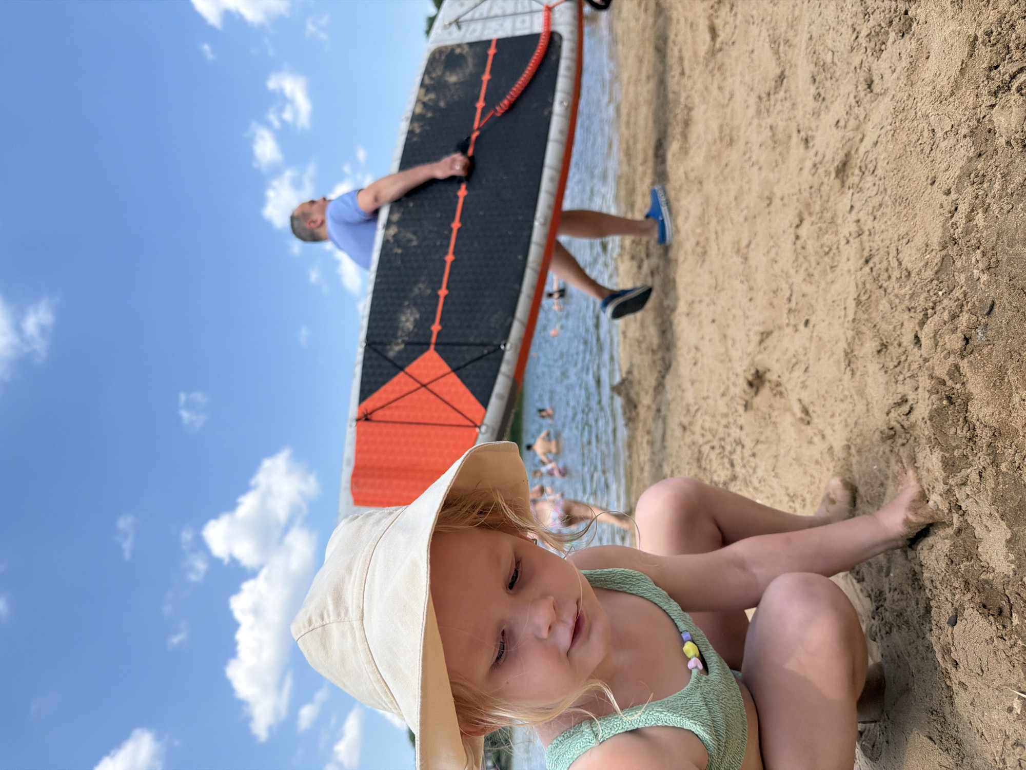The media depicts a sunny day at the beach, likely in Malakhovka, Russia. In the foreground, a young child wearing a light-colored bucket hat and a green swimsuit sits on the sandy shore, with their legs crossed. The child is adorned with a colorful beaded necklace. Behind the child, a man is walking on the sand, carrying a large paddleboard. The paddleboard has a black and orange design. The water is visible in the background with several other people swimming or wading. The sky is blue with scattered white clouds. The overall scene suggests a relaxed summer day at a lakeside or beach.