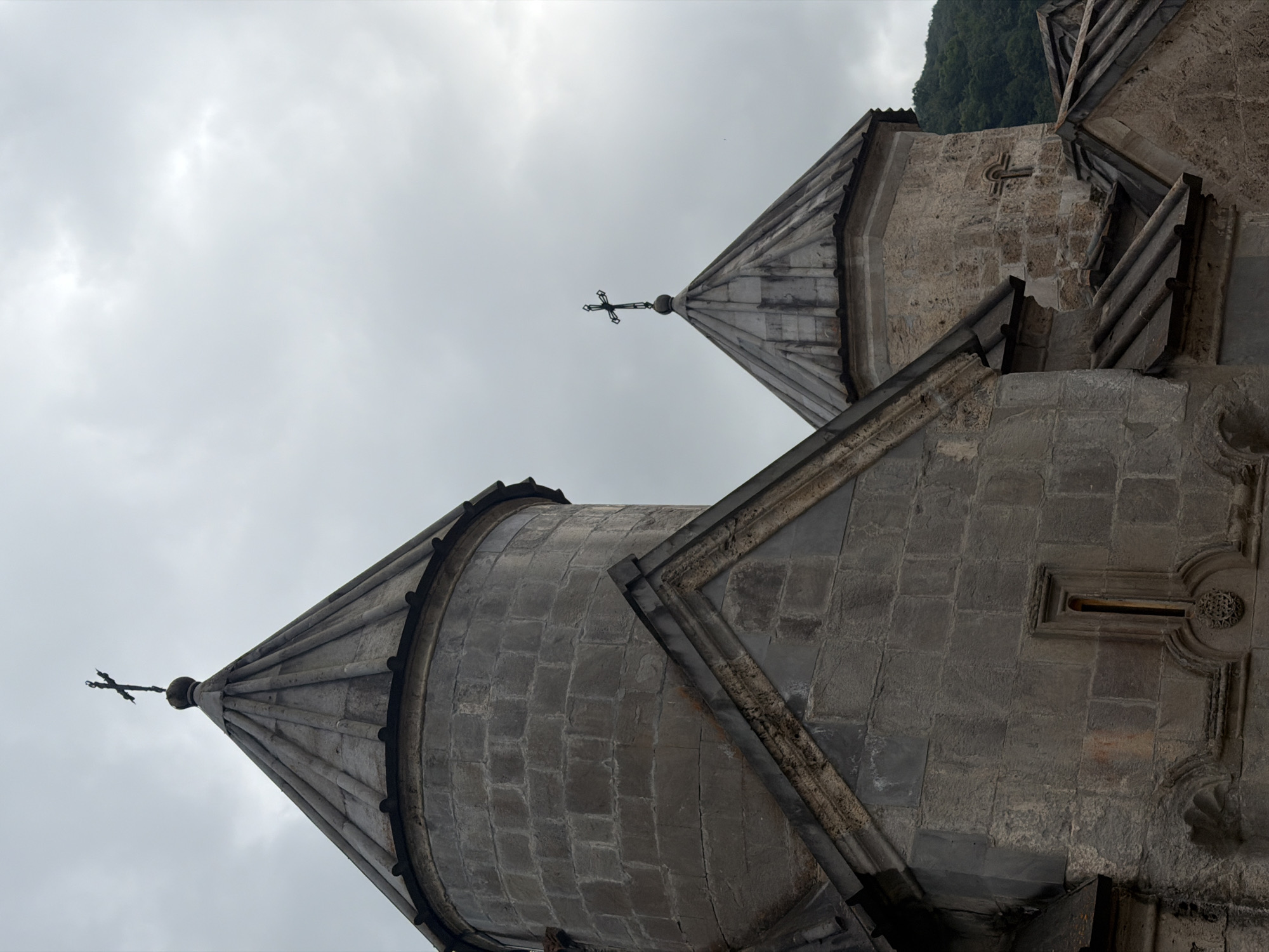The image depicts a low-angle view of two conical stone roofs of ancient religious structures, possibly churches or monasteries. The weathered stone blocks forming the roofs are visible, with one roof topped by a dark cross and the other featuring a more ornate carved cross or symbol integrated into its structure. The background is a cloudy, overcast sky, suggesting a temperate or cool climate. To the upper right, a densely forested mountainside is visible, indicating a rural or mountainous location. The architecture suggests a historical and possibly religious setting, consistent with the known location context of Teghut, Armenia, which is known for its medieval churches. No people or specific activities are depicted. The scene evokes a sense of historical depth and solemnity due to the architecture and the muted lighting. There is no visible text in the image.