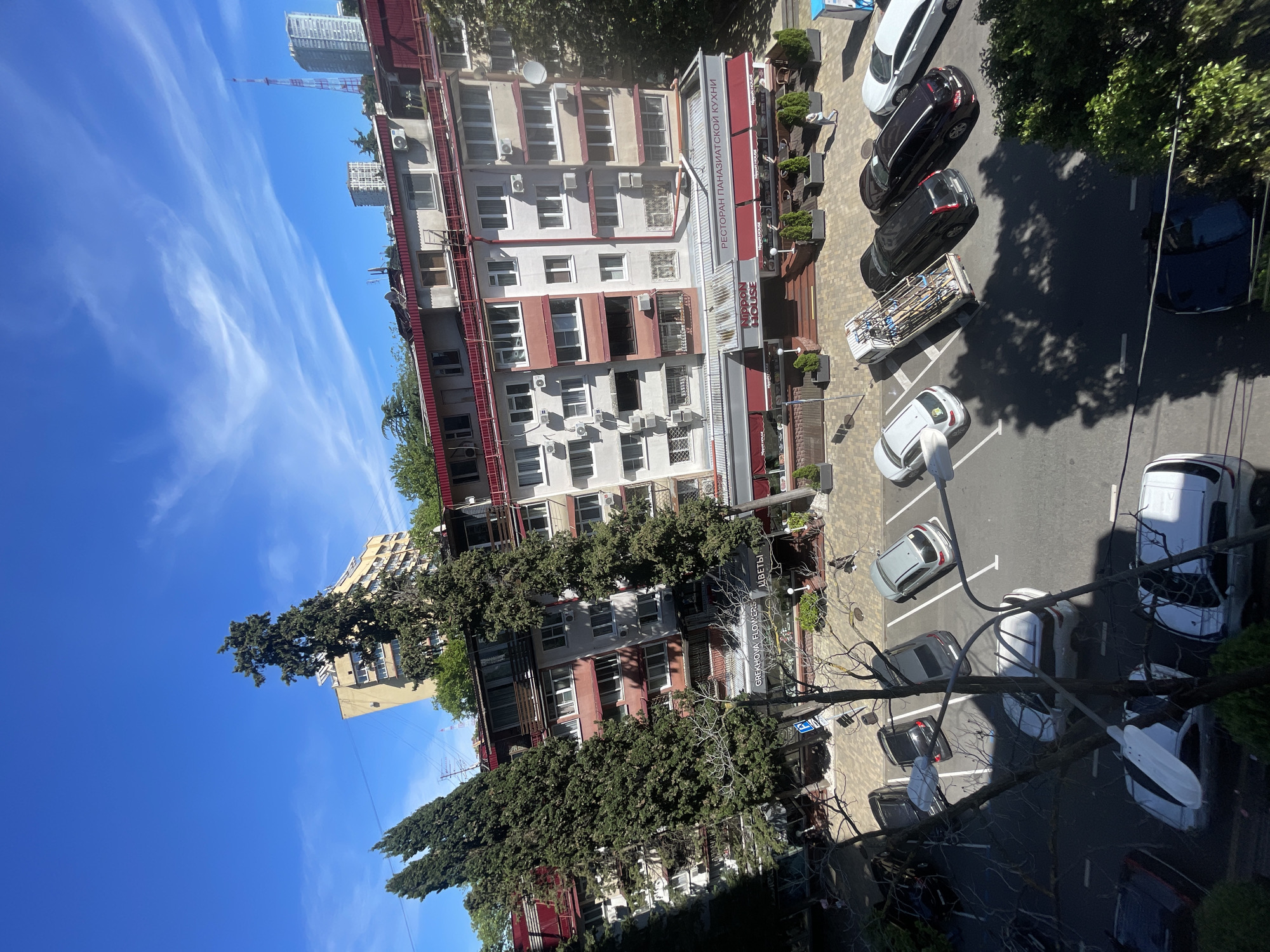 The image presents an elevated view, likely from a window or balcony, looking down upon a bustling street scene in a city, identified as Sochi, Russia. The perspective is tilted as if the camera was held sideways, but the description assumes a corrected upright orientation.
In the foreground and midground, a multi-story building dominates, featuring several residential floors with balconies and windows. The ground floor houses commercial establishments with prominent red awnings and signage. Numerous air conditioning units are visible on the building's facade. Along the street in front of this building, several cars are parked, including a white truck with a large metal rack on its bed.
Further down the street, more cars are parked in what appears to be a parking lot area, partially shaded by large, mature trees. Streetlights are also visible along the curb. To the left of the main building, another distinct yellow-colored building with a large evergreen tree in front of it can be seen. In the distance, taller modern buildings and a telecommunication tower pierce the horizon.
The sky is a vibrant clear blue with wispy white clouds, indicating bright, sunny weather. The strong shadows cast by the buildings and trees suggest it is midday or early afternoon. No explicit human activity is clearly visible, making the scene appear relatively quiet, focusing on the urban infrastructure.
Visible text on the main building's awnings and signs includes Cyrillic script: "РЕСТОРАН ПАНАЗИАТСКОЙ КУХНИ" (Restaurant Pan-Asian Cuisine) and "NOON HOUSE". Another storefront displays "GREKHOVA FLOWERS ЦВЕТЫ" (Grekova Flowers Flowers). A blue parking sign with a white "P" is also visible on the street.