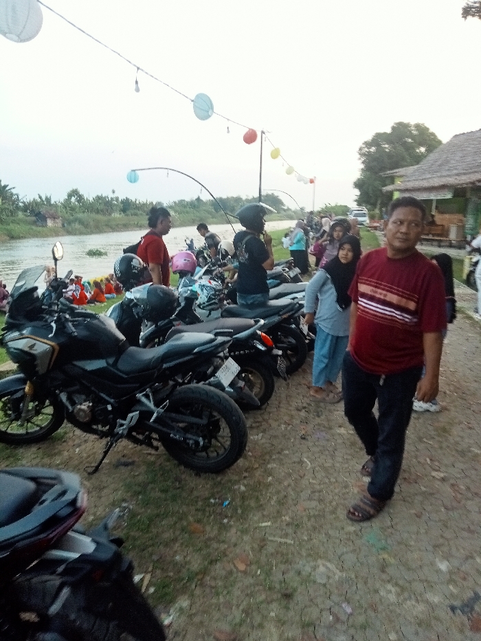 The scene takes place in Jakenan, Indonesia, at the coordinates -6.758 latitude and 111.075 longitude. It is a gathering of people, mostly men, by the river. There are several motorcycles parked along the riverbank, some with helmets resting on them. A man wearing a red shirt with white stripes and black pants stands in the foreground, looking toward the right. A woman in a light blue headscarf and a long blue dress stands to his left, with a woman in a pink headscarf to her left. There are other people standing in the background, including a man in a blue shirt and a woman in a pink shirt. There is a long fishing pole lying on the ground, and a row of colorful paper lanterns hang from a string above the scene. The ground is covered in dirt and grass. The atmosphere is casual and relaxed. There appears to be a grassy area, trees and buildings in the background. The river is visible in the background. The sky is cloudy and overcast.