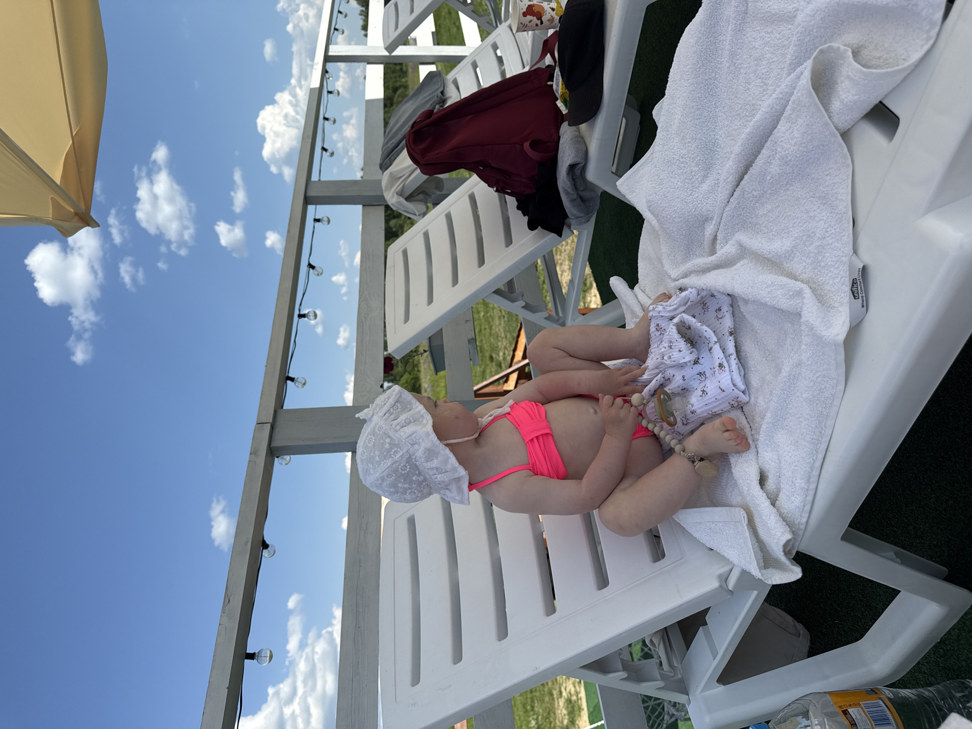 A baby wearing a pink bikini and a white sun hat lies on a white plastic lounge chair covered with a white towel. The baby is on their back, with legs bent and feet facing upwards. A white cloth with a floral pattern is tucked under the baby's legs. A pacifier with a beaded string is attached to the baby's hand. In the background, there are other empty lounge chairs, some with clothing draped over them, and a wooden structure with string lights. The scene appears to be outdoors, possibly at a resort or recreational area, with a clear blue sky and white clouds visible above. The lighting suggests it is daytime. There is no visible text in the image.