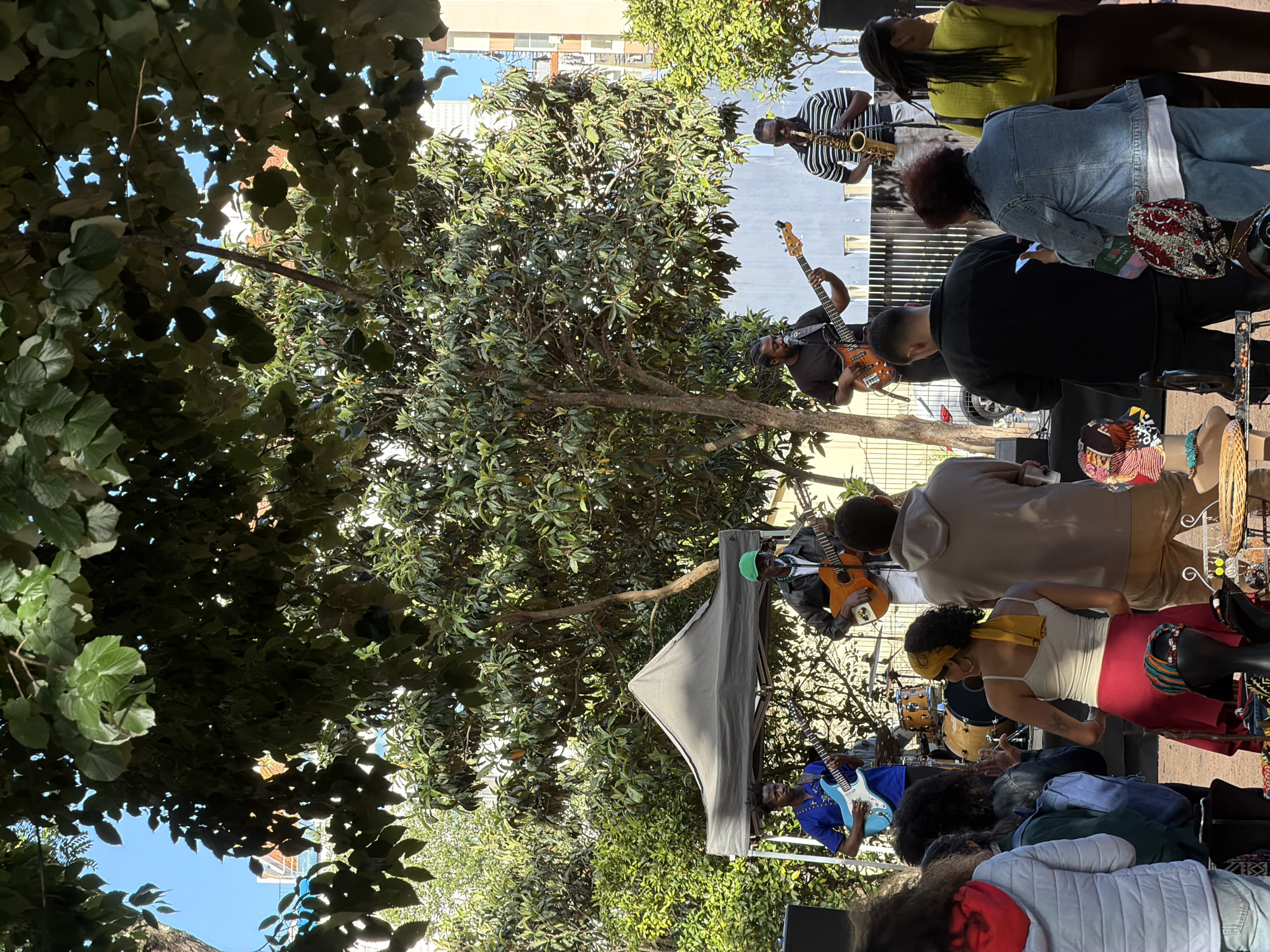 The image shows an outdoor music performance in what appears to be a public space in Lisbon, Portugal. A band composed of a saxophone player, a bass guitarist, a drummer, and two guitarists, is performing. The musicians are of African descent, and a tent is set up, possibly to provide shade or as part of the setup. An audience, comprised of people of varying ages and ethnicities, is watching the performance. Some members of the audience are standing while others are seated. Merchandise, possibly related to the band or the event, is displayed on a table next to the audience. The setting includes trees and buildings, suggesting the event is taking place in a park or square within the city.