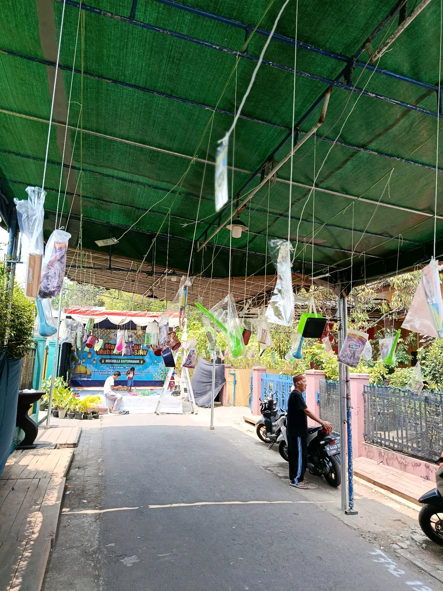 The image depicts an outdoor scene in Bululawang, Indonesia, likely a community event. A green tarp stretches across the top of the frame, supported by poles and strung with numerous items encased in clear plastic bags, suspended by white strings. These items appear to be prizes or decorations, some resembling toys or household goods. Below the tarp, a paved walkway runs alongside a road. In the background, a banner is set up with text that appears to be in Indonesian, possibly announcing an event for "Musholla Baiturrahman". Two individuals, a man in a white shirt and pants, and a child in a blue shirt and shorts, are seen near the banner, seemingly engaged in an activity on a low platform. To the right of the walkway, a man in a black shirt and pants, wearing a red cap, stands next to a motorcycle. The environment includes potted plants and fencing, suggesting a residential or communal area. The overall atmosphere is bright and sunny, indicating daytime.