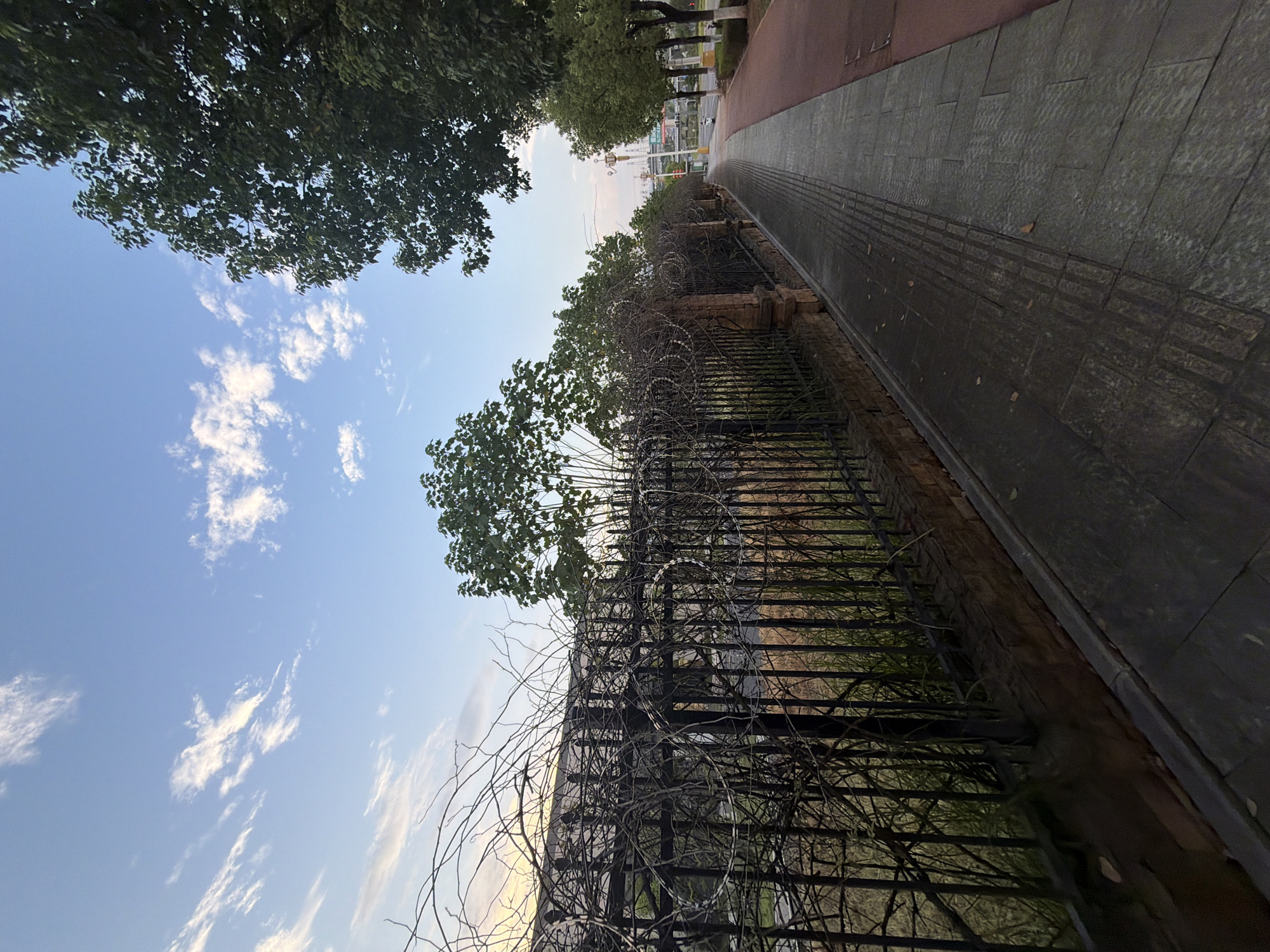The image depicts a sidewalk with a black metal fence on the left, topped with coiled wire. The fence is partially obscured by bare branches and some green foliage. Beyond the fence, dry grass and vegetation are visible. A paved walkway runs alongside the fence, and to the right, a reddish-colored path is present. Trees line the right side of the reddish path, and in the distance, buildings and infrastructure are discernible, including a tall structure with lights. The sky is a pale blue with scattered white clouds. The scene appears to be set in an urban or semi-urban environment, possibly a park or street, in Jiangjin, China. No people are clearly visible in the image.