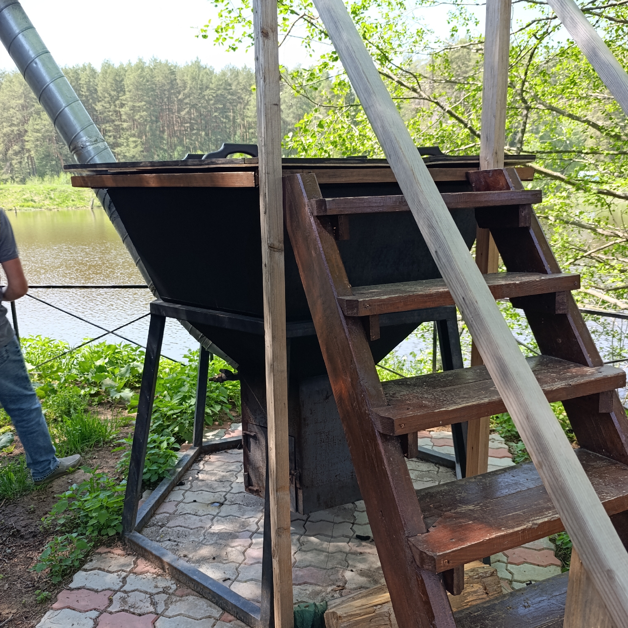 The image depicts an outdoor scene in Prikamskii, Russia, featuring a large, custom-built cooking apparatus. This structure consists of a conical black cooking pot mounted on a metal frame with a chimney extending upwards. A wooden staircase provides access to the top of the cooking pot. The apparatus is positioned near a body of water, likely a river or lake, bordered by vegetation. A man is partially visible on the left side of the frame, standing near the water's edge. The ground around the apparatus is paved with patterned tiles. The surrounding environment includes trees and greenery, suggesting a natural or park-like setting.