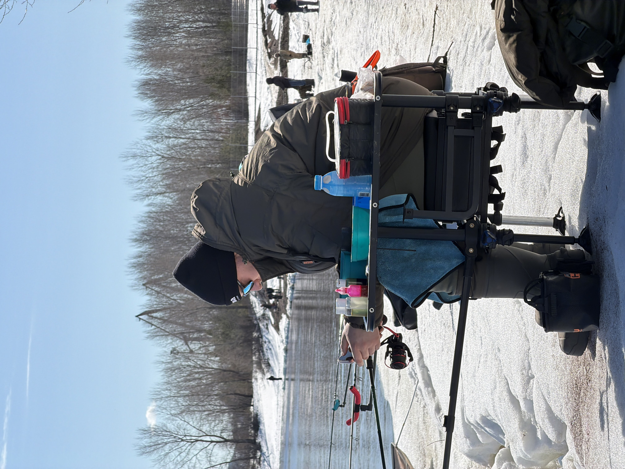 A person wearing a black beanie, sunglasses, and a green jacket is fishing on a frozen body of water. The person is sitting on a fishing chair with various attachments, including a water bottle and small containers. Multiple fishing rods are set up. The ground is covered in snow. In the background, bare trees line the horizon under a clear blue sky. Several other people are visible in the distance, also appearing to be fishing. The location is the city of Chaikovsky, Russia.