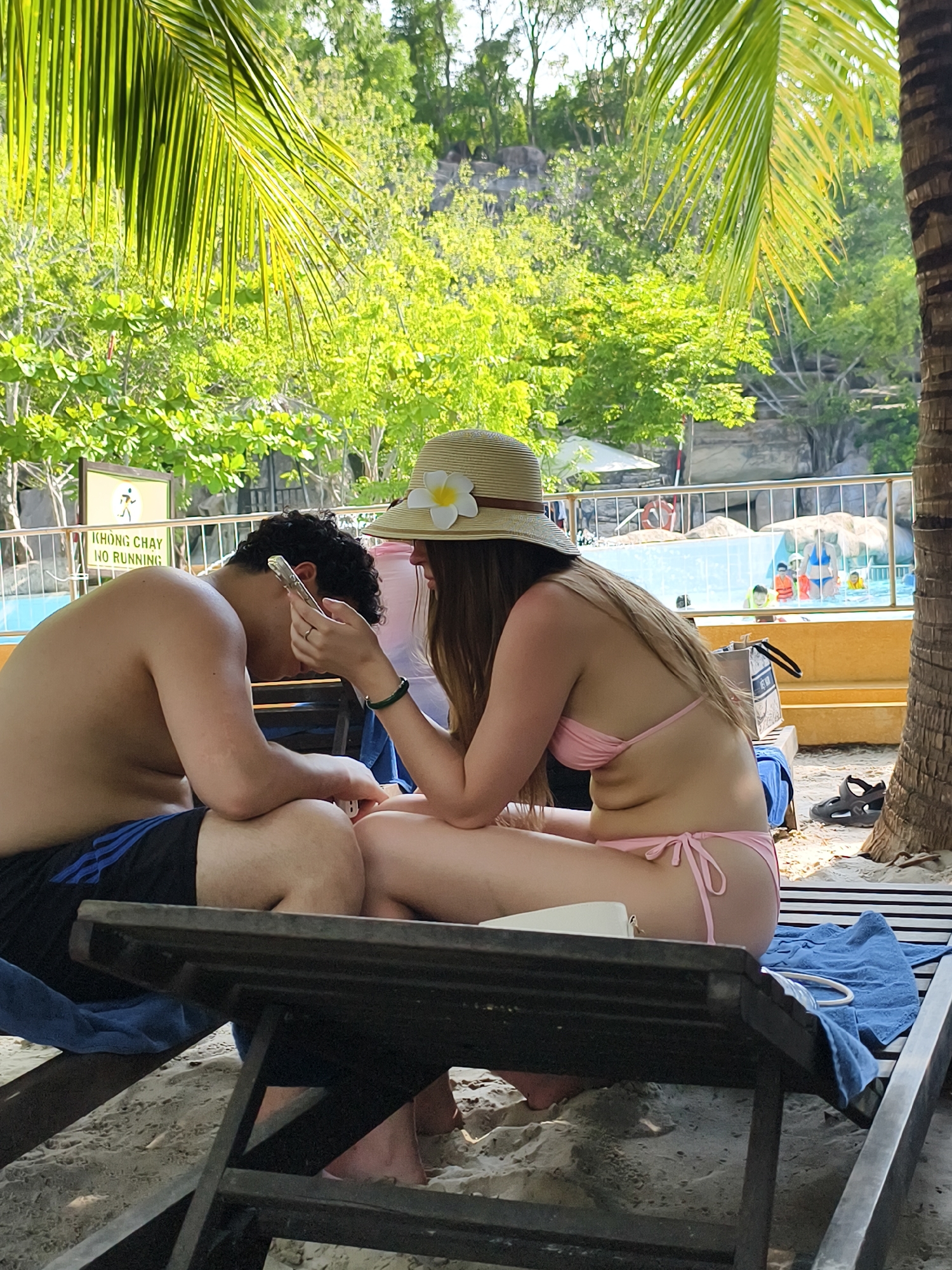 A woman in a pink bikini and straw hat with a flower is seated on a beach chair next to a man in black shorts. The man is holding and looking at a smartphone. They are situated on a sandy beach, under palm trees, near a pool. A sign that reads "KHONG CHAY NO RUNNING" is nearby. In the background, people are in the pool. The location is Unknown, Unknown.
