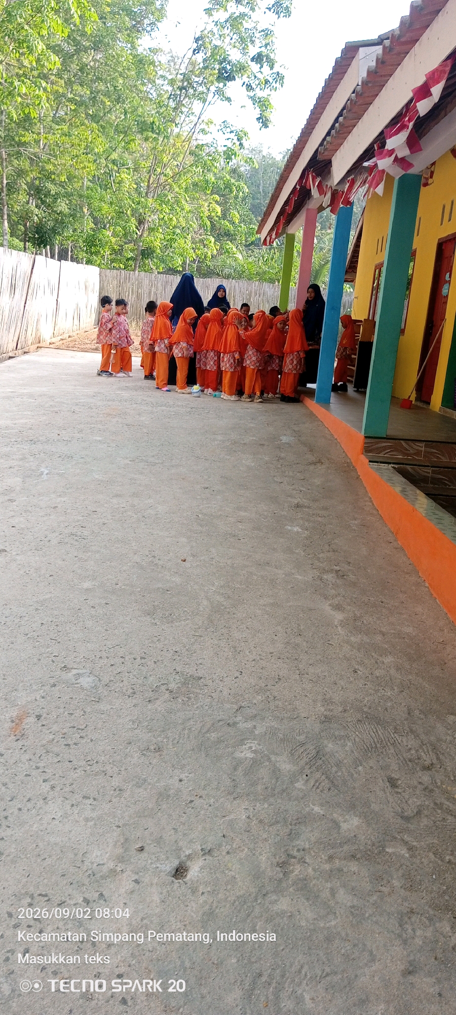 A group of young children, some wearing orange hijabs and matching outfits, stand in a line on a concrete path. Two women, one in a dark blue hijab and the other in a lighter hijab, stand behind the children. The children are facing towards a building with a yellow facade and a red tiled roof decorated with Indonesian flags. The building has colorful pillars supporting an overhang. A wooden fence is visible to the left of the scene, behind which are trees and foliage. The image is watermarked with "2026/09/02 08:04 Kecamatan Simpang Pematang, Indonesia Masukkan teks" and "TECNO SPARK 20".