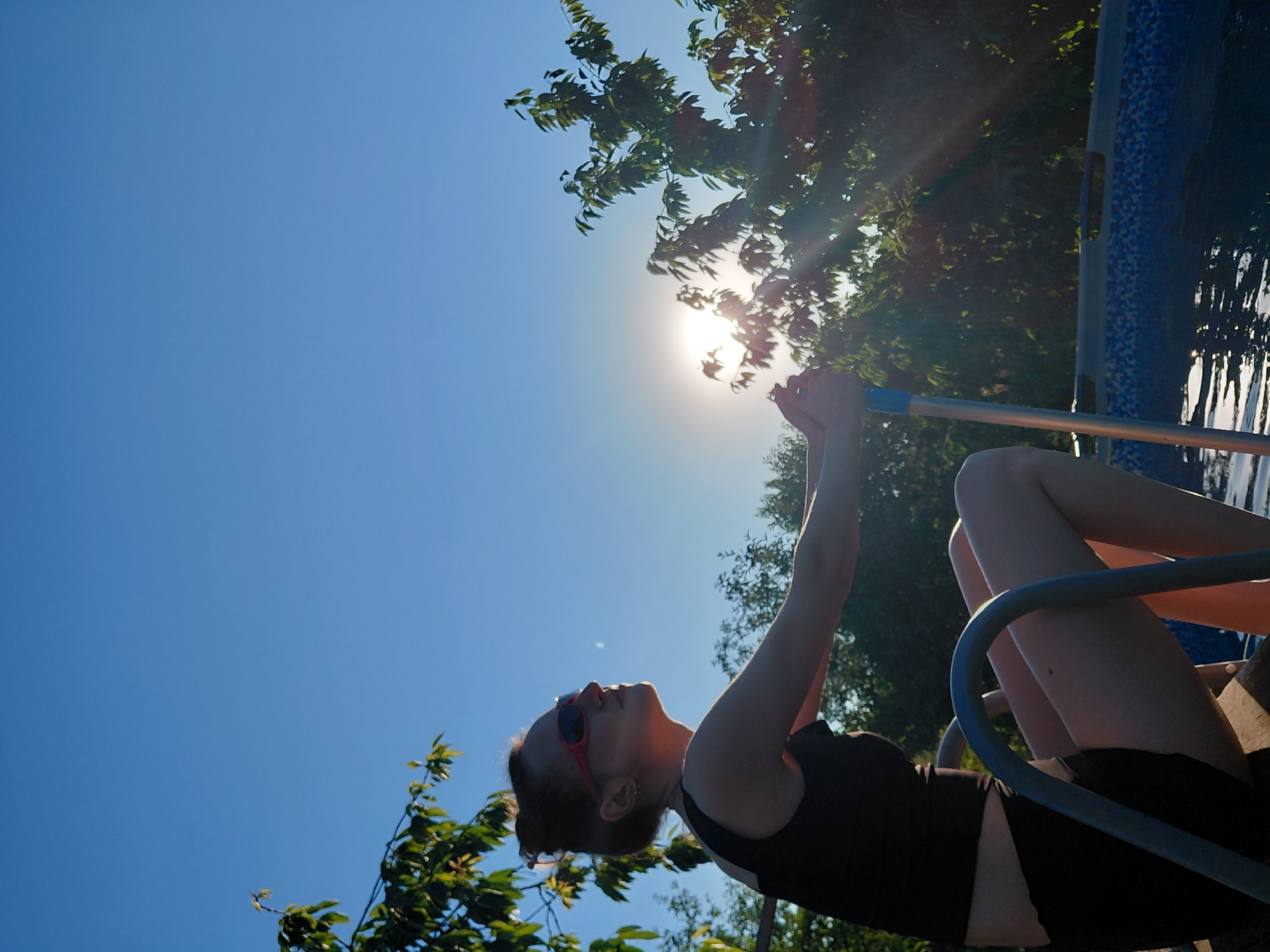 A low-angle, rotated shot shows a woman in a black swimsuit sitting in a chair outdoors. She is wearing red-rimmed sunglasses. Her arms are raised and outstretched, and she is holding a silver pole with a blue handle. Sunlight streams down from the top right, creating lens flare and highlighting her form. In the background, there is a blue and white patterned pool. The scene is framed by green foliage, suggesting a sunny day. The overall impression is one of leisure and enjoyment. The image is tilted, giving a dynamic perspective to the scene. The time of day appears to be late afternoon or early evening, indicated by the low angle of the sun.