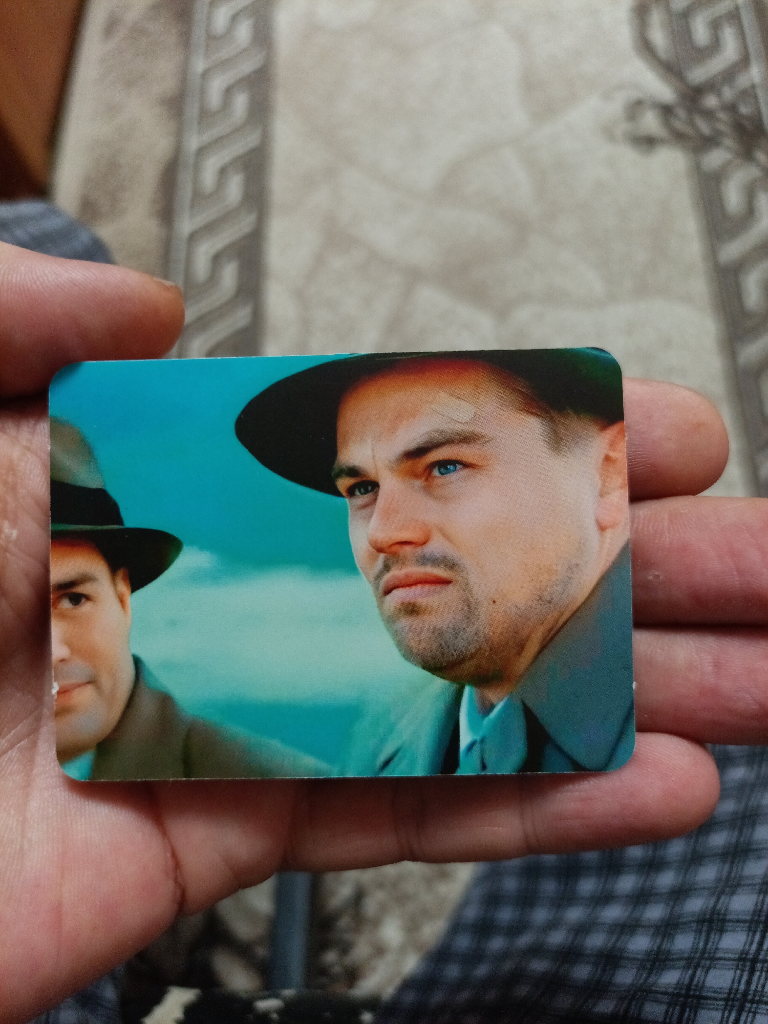 An image shows a close-up of a postcard or trading card featuring Leonardo DiCaprio and another man wearing hats. The card is held in a person's hand, with portions of the hand visible. The background of the card depicts a bright blue sky. There is no indication of the city of Nyagan, Russia in the image.