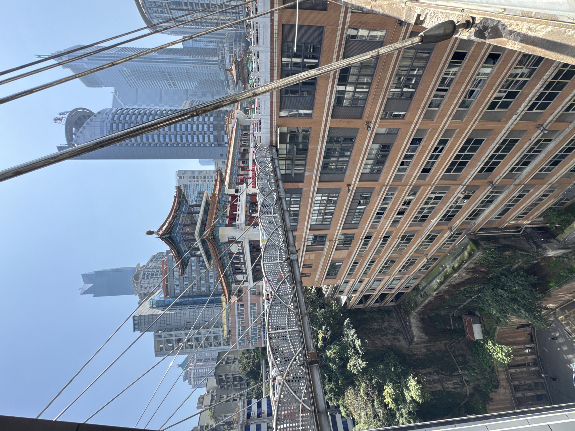 The image shows a view in Yuzhong District, China. A pedestrian bridge with a decorative lattice design spans across a green space between buildings. On one side of the bridge are modern skyscrapers, and on the other side is a multi-story building with a brick facade and numerous windows. The bridge is suspended by steel cables. A traditional Chinese-style structure with a tiered roof is also visible amongst the buildings. The sky is clear and blue.
