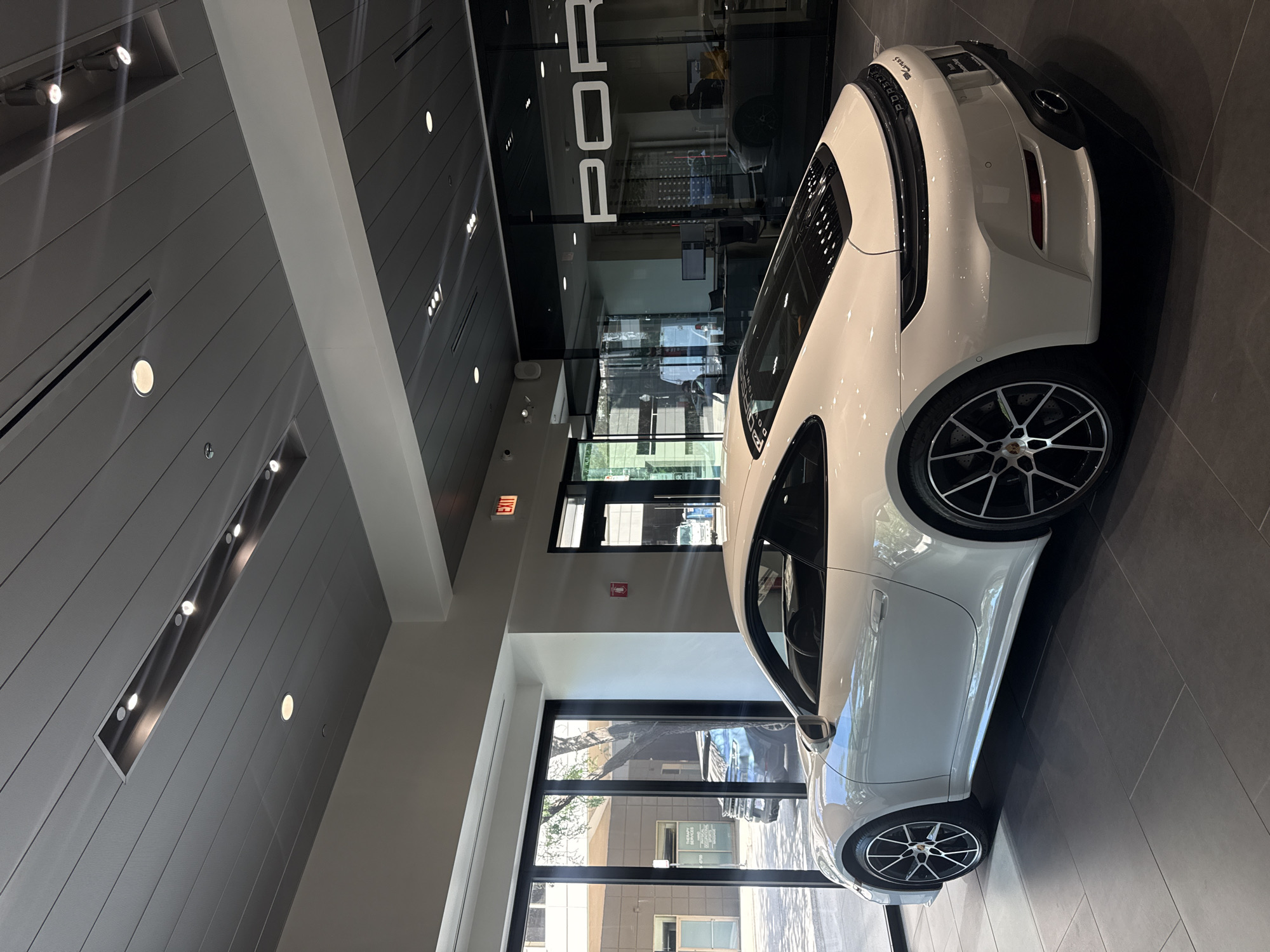 A white Porsche 911 Carrera S is parked indoors on a tiled floor in a showroom. The car is viewed from its rear right quarter and is facing left. The showroom has a modern design with a suspended ceiling featuring recessed lighting and linear vents. Through a large glass window on the left, an outdoor scene is visible, showing a street, trees, and buildings. The window on the right has the "PORSCHE" logo displayed. An exit sign is illuminated in red above a doorway. The car's details include its distinctive taillight bar, quad exhaust pipes, and black alloy wheels with silver brake calipers. The interior appears clean and well-lit, typical of a luxury car dealership. There are no people clearly visible in the immediate vicinity of the car, although reflections in the glass suggest some activity outside.