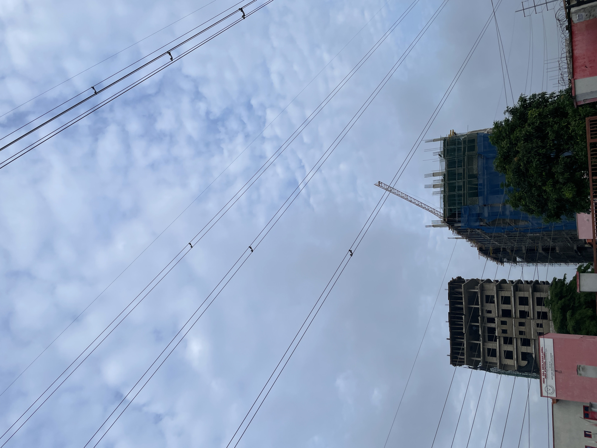 The image shows an overcast sky with numerous power lines crisscrossing the frame. In the middle ground, two unfinished buildings are visible. The building on the left is a concrete structure with numerous window openings. The building on the right is partially covered with blue netting and has a crane extending from its top. A large green tree is situated in front of the building on the right. In the foreground, a pink building with a sign reading "MRM OBA GBOLAHAN LAWAL FIRE STATION AREA" is partially visible. The image appears to be taken in the city of Itirin, Nigeria.
