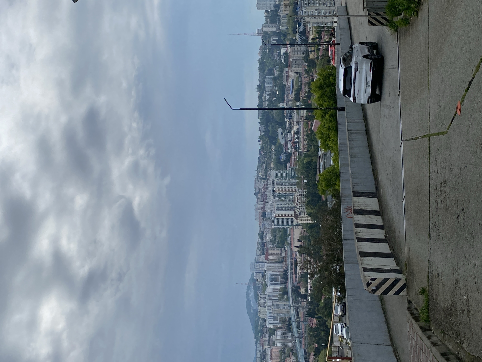 This photograph depicts a cityscape of Sochi, Russia under a cloudy sky. The vantage point is elevated, likely from a road or viewpoint overlooking the city. The city is dense with buildings of varying heights, many appearing to be apartment complexes. A tall communications tower is visible in the middle ground. In the foreground, a concrete barrier with black and white striped markings lines the right side of the frame, next to a road with a white car parked on it. Additional cars are seen parked further down the road. Streetlights are also present. The overall environment is urban with some greenery interspersed among the buildings.