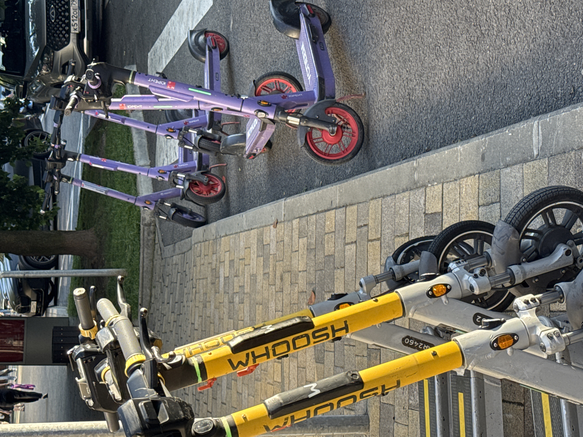In Sochi, Russia, several parked electric scooters are visible. Two yellow scooters with "WHOOSH" branding are in the foreground, positioned on a textured paving surface. Behind them, several purple electric scooters with Cyrillic branding are lined up on an asphalt surface, some with red wheels. A dark-colored SUV with a license plate visible is parked in the background, along with a tree. A person wearing dark clothing and carrying a purple bag is walking away from the camera in the upper left corner of the image. The scene appears to be a street or sidewalk area.