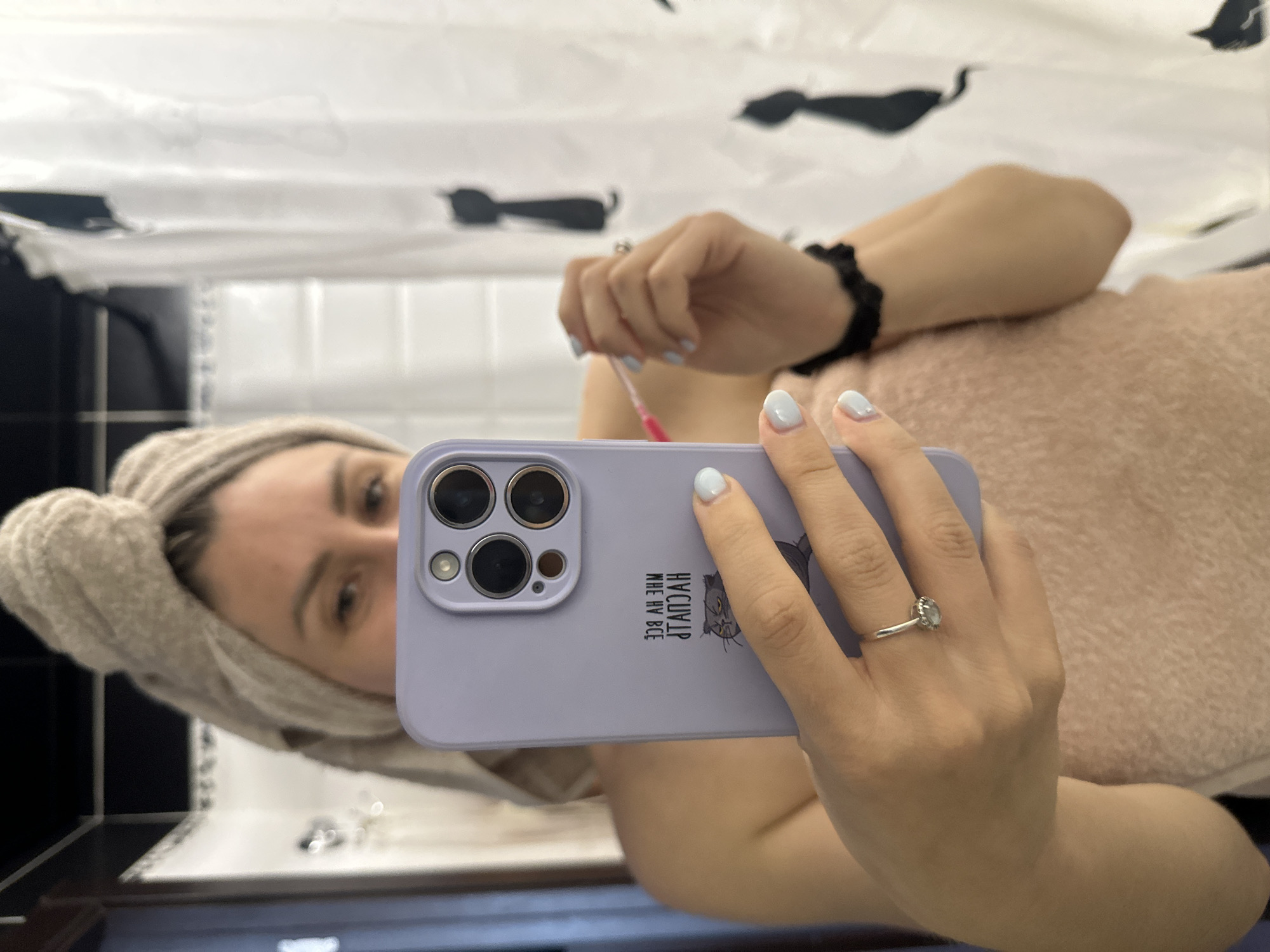 A woman with a towel wrapped around her head is taking a selfie in a bathroom. She is holding a purple phone in her left hand, which has a camera lens cover and a graphic of a cat. She is wearing a ring on her left ring finger. In her right hand, she is holding a pink cotton swab near her face. A light pink towel is wrapped around her torso. The background shows a shower curtain with black cat silhouettes. The scene appears to be in a residential bathroom.