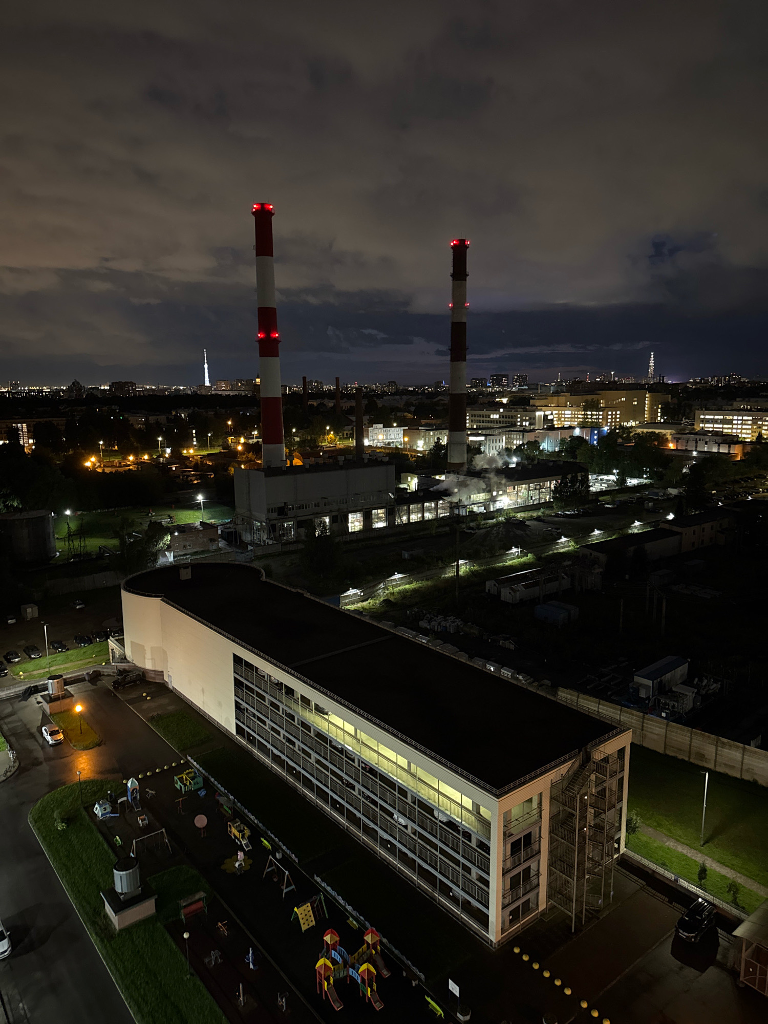 This image, taken at night from a high vantage point, depicts an industrial area within Saint Petersburg, Russia, likely the Kirovsky District. Dominating the skyline are two tall, red and white striped industrial chimneys, emitting plumes of steam. In the foreground, a long, multi-story building with numerous windows is visible. Adjacent to this building is a playground with various colorful structures. The surrounding landscape is a mix of industrial facilities, residential buildings, and streetlights illuminating the dark urban environment. The sky is cloudy, with some of the clouds illuminated by city lights. No individuals are clearly discernible in the image.
