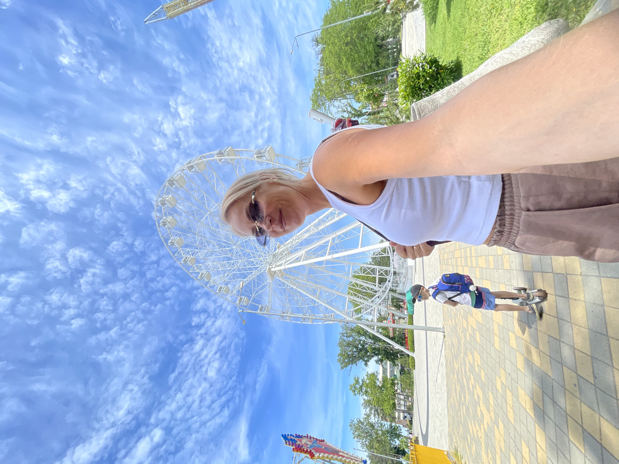 A blonde woman wearing a white tank top and brown shorts takes a selfie in front of a Ferris wheel. The woman is wearing sunglasses and has her arm extended to capture the photo. In the background, a young boy in a blue backpack and a cap is riding a scooter on a patterned pavement. The Ferris wheel is white and large, dominating the background. There are green trees and a bright blue sky with white clouds. The location appears to be an amusement park or recreational area in Anapa, Russia.