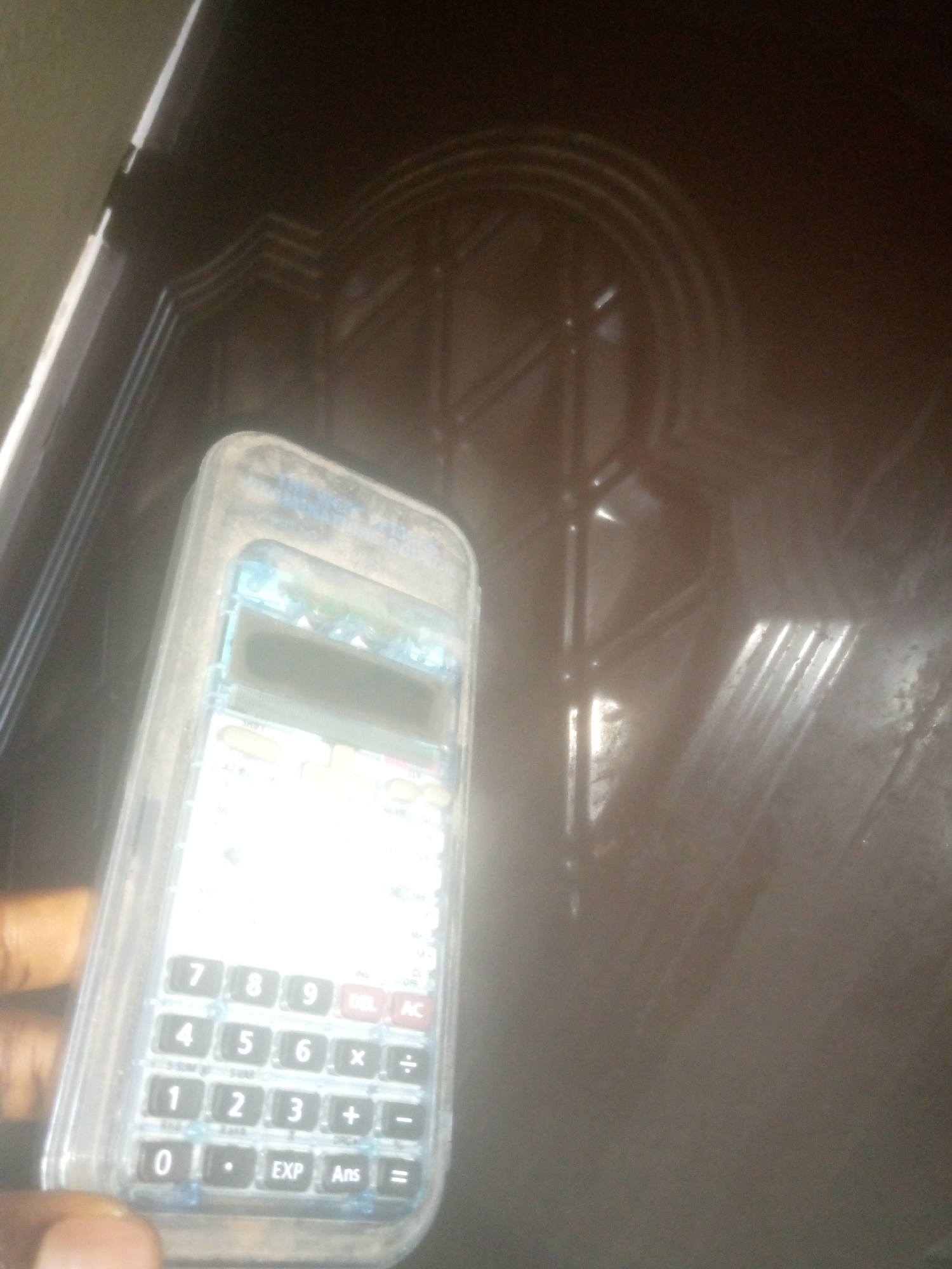 A hand holds a clear plastic-cased calculator with a white keypad against a dark, textured surface, possibly a door. The calculator has standard number keys (0-9), arithmetic operators (+, -, x, /), a decimal point, EXP, and Ans buttons. The display screen is visible but unreadable due to glare. The dark surface behind the calculator features a raised, geometric pattern, with a circular motif in the upper right portion of the frame. The lighting is dim, creating reflections on both the calculator and the background surface.