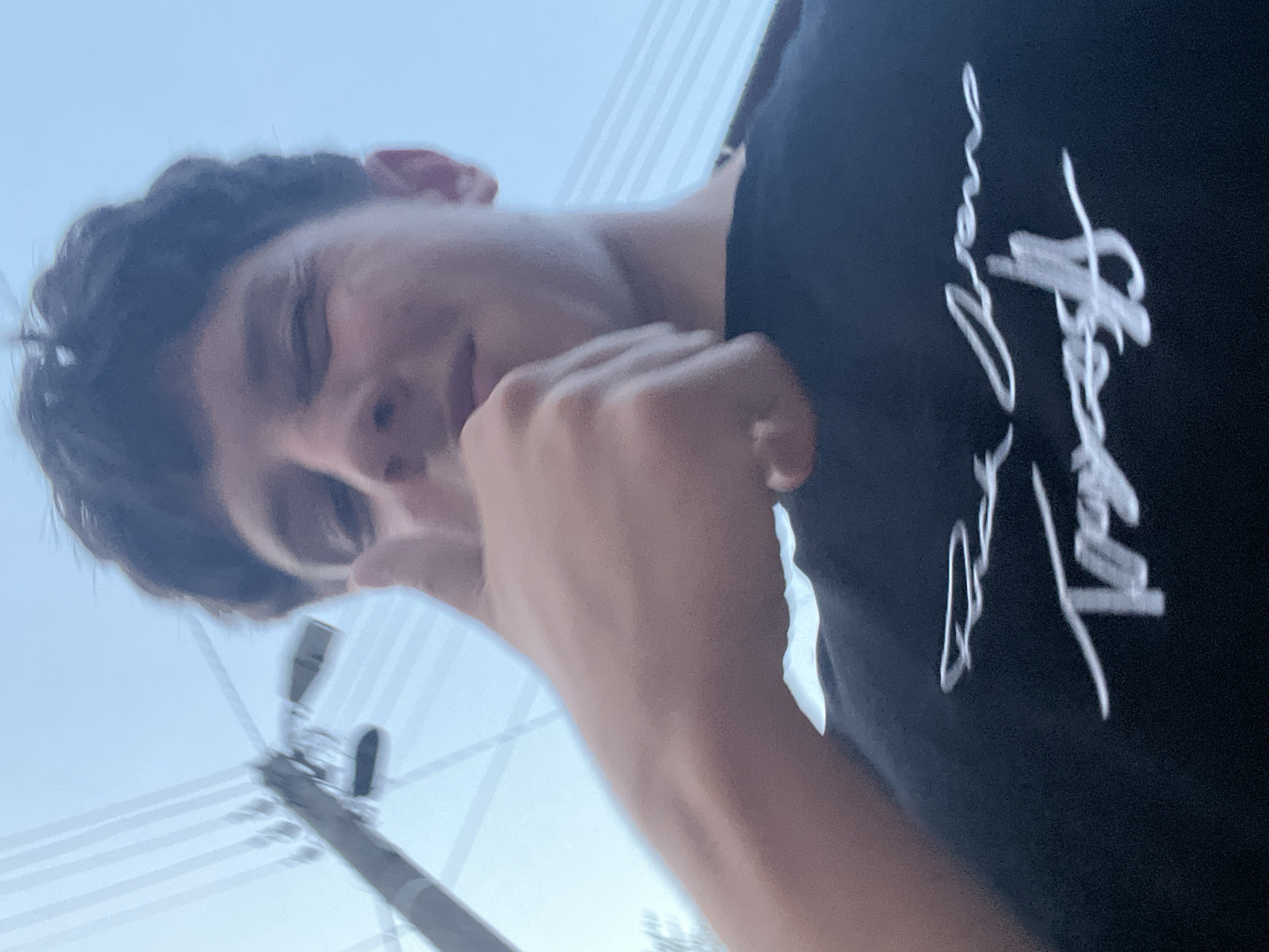 The image depicts a young male, possibly adolescent, positioned in a low-angle, close-up shot against a light blue sky, in Zlatograd, Bulgaria. He has dark, wavy hair and is looking towards the camera, with his left eye partially closed as if winking or squinting. His right arm is raised, and his hand forms a thumbs-up gesture, with the thumb extended vertically and the other fingers curled into a fist, directed towards the viewer. He is wearing a dark, likely black, t-shirt with white, stylized text or a signature embroidered or printed on the front. In the upper left background, a blurred utility pole with multiple horizontal wires is visible against the clear sky. The scene is outdoors and brightly illuminated by natural light.