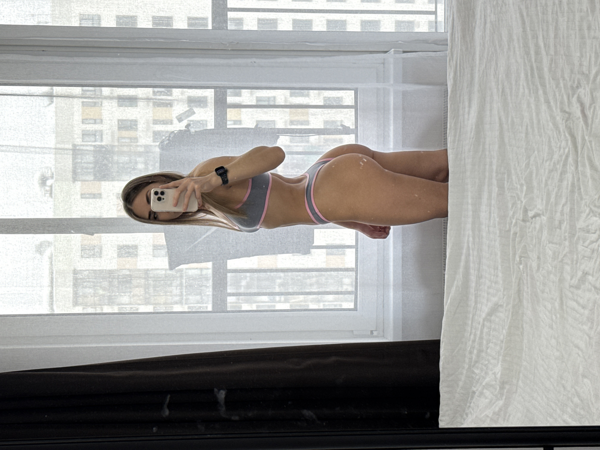 A woman is taking a mirror selfie in an apartment, wearing a gray and pink sports bra and matching underwear. The cityscape visible through the window suggests a location in Lyubertsy, Russia, based on the architecture. The scene includes a white sheet partially covering the right side of the frame. A phone is used to take the picture.