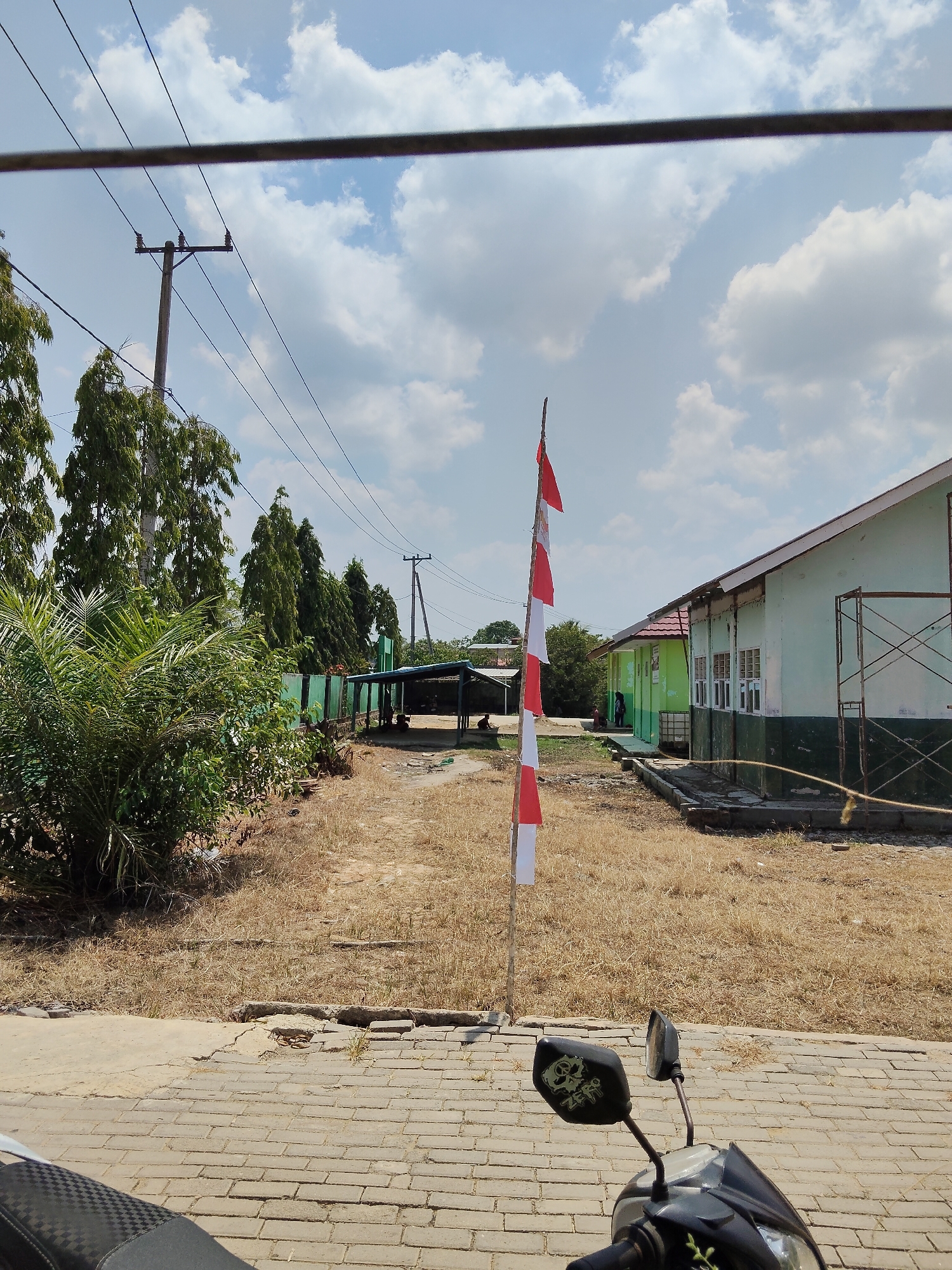 The image depicts a scene outside a building in Lori, Indonesia. In the foreground, a motorcycle is partially visible, with its handlebars and mirrors facing the viewer. A red and white flag, likely the Indonesian flag, is planted in the ground to the left of the motorcycle, in front of a building. The building itself appears to be light green with a darker green base and a red roof. Scaffolding is erected on the right side of the building.
The surrounding environment is dry and somewhat unkempt. A pathway made of brick pavers is in the foreground, transitioning to a dry, grassy area. Tall trees with dense foliage line the left side of the scene, and more trees are visible in the background. Electrical poles and wires are prominent in the upper portion of the image, spanning across the sky. The sky is blue with scattered white clouds, suggesting a sunny day. In the middle ground, there is a covered open-air structure, possibly a waiting area or shelter, with a few people sitting or standing beneath it.