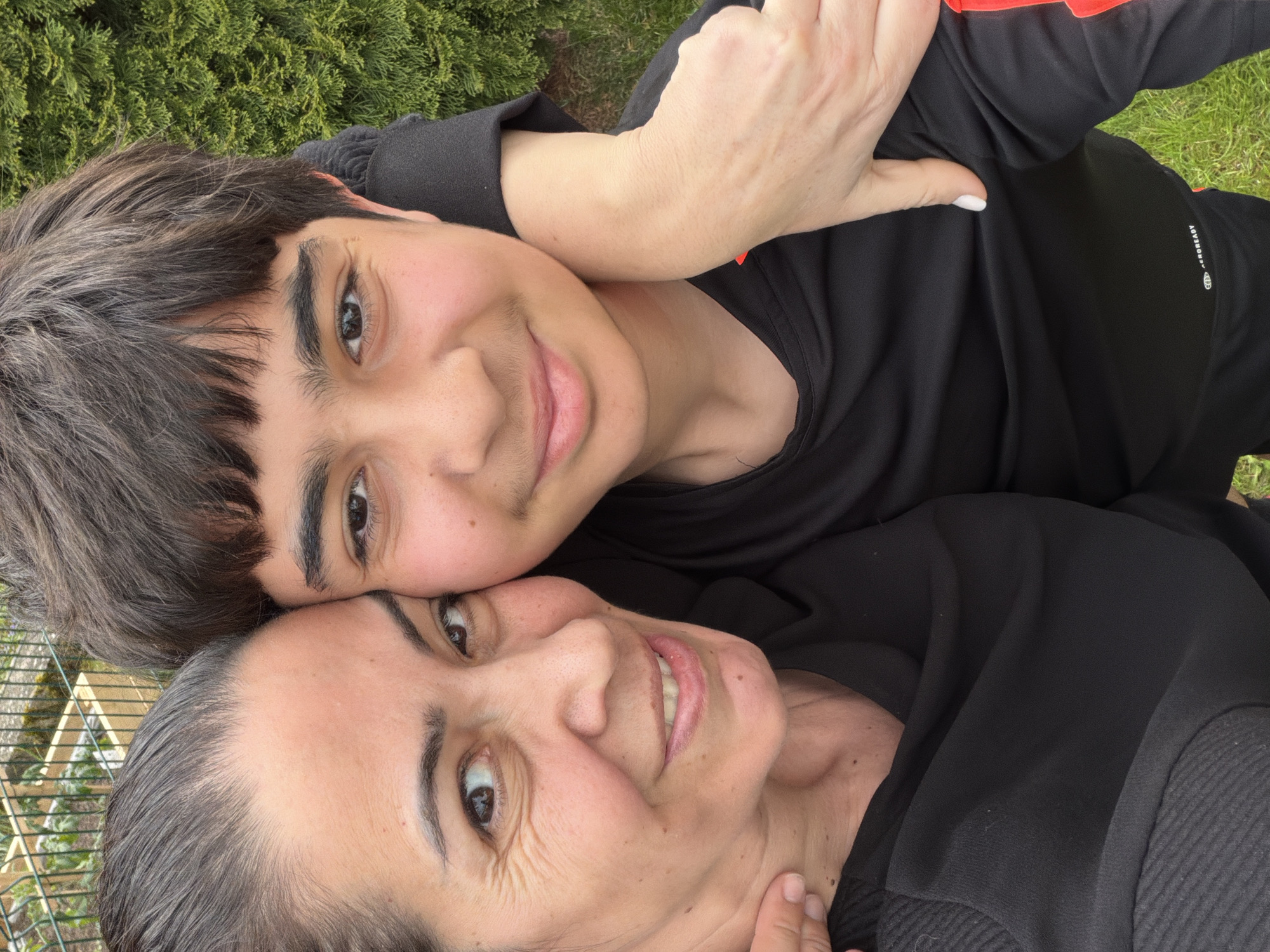 Two people are lying on the ground, likely grass, in an outdoor environment, possibly in Esch-sur-Alzette, Luxembourg. The person on top appears to be younger, with dark hair and wearing a black athletic shirt with red stripes on the sleeves. The person below appears to be older, with dark hair, and also wearing a black shirt. They are close together, possibly embracing or in a selfie pose. Green foliage is visible in the background and there is what appears to be a fence with foliage behind it.