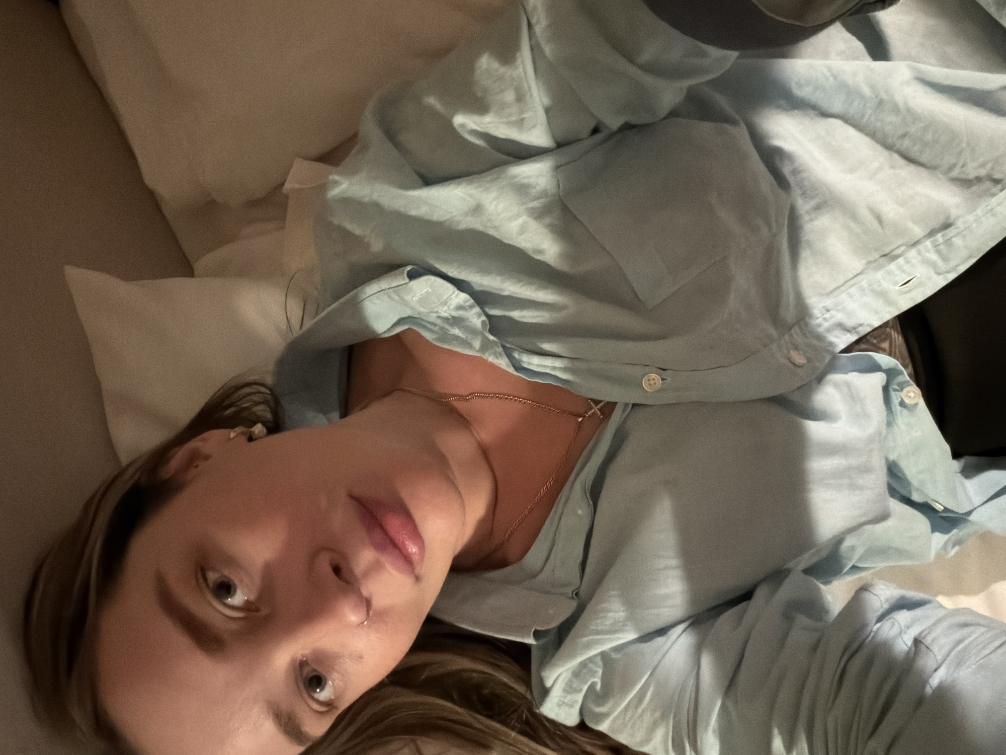 A woman lies on a light-colored pillow, wearing a light blue, collared shirt with a cross necklace. Her hair is light brown, and she is looking at the camera. The background includes a light-colored surface, likely a bed or couch. The location is not evident from the image.