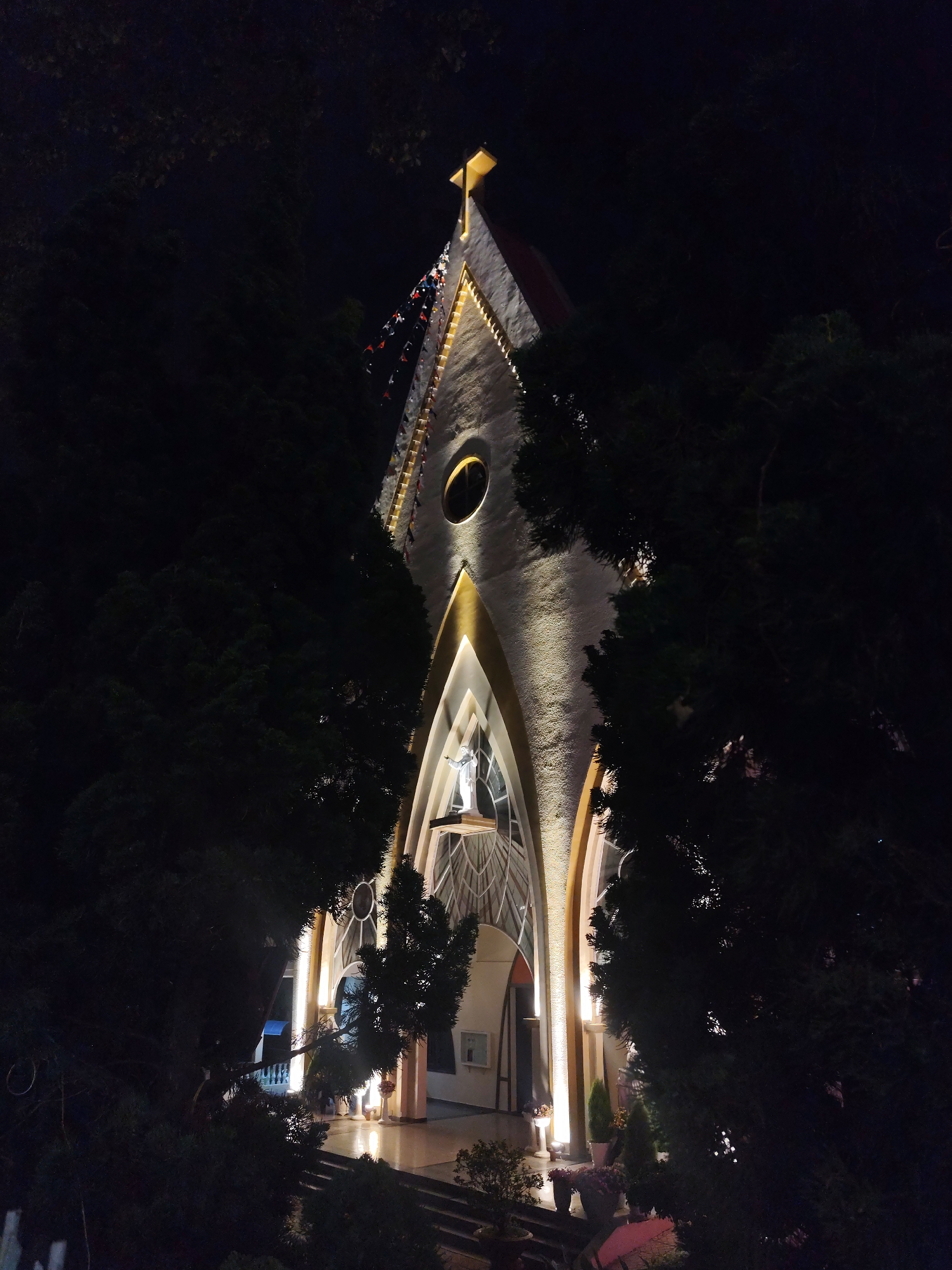 The image shows the Domaine de Marie Church in Da Lat, Vietnam, at night. The church is illuminated with golden lights outlining its architectural features. A statue is visible within the entrance archway. The church is framed by dark trees on both sides. The image has a low-light, nighttime atmosphere. Decorations are hung along the roofline. A cross sits on top of the steeple. Steps lead up to the church entrance. Flowers in pots are placed on the steps.