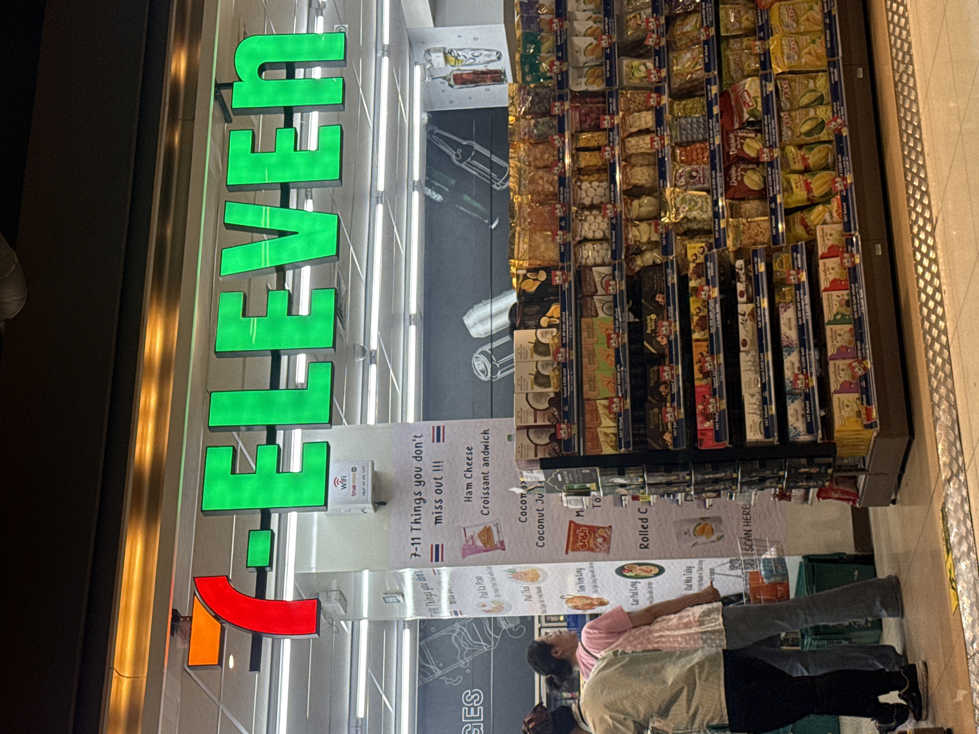 The image shows the exterior of a 7-Eleven convenience store in Nong Prue Subdistrict, Thailand. The iconic red and green 7-Eleven logo is prominently displayed above the entrance. Inside the store, shelves are stocked with various food items, including snacks, drinks, and what appear to be packaged meals. A few customers are visible, browsing the merchandise. The lighting suggests it is daytime, and the store appears to be clean and well-organized. A promotional banner inside the store lists "7-11 Things you don't miss out!!!" with images of food items.