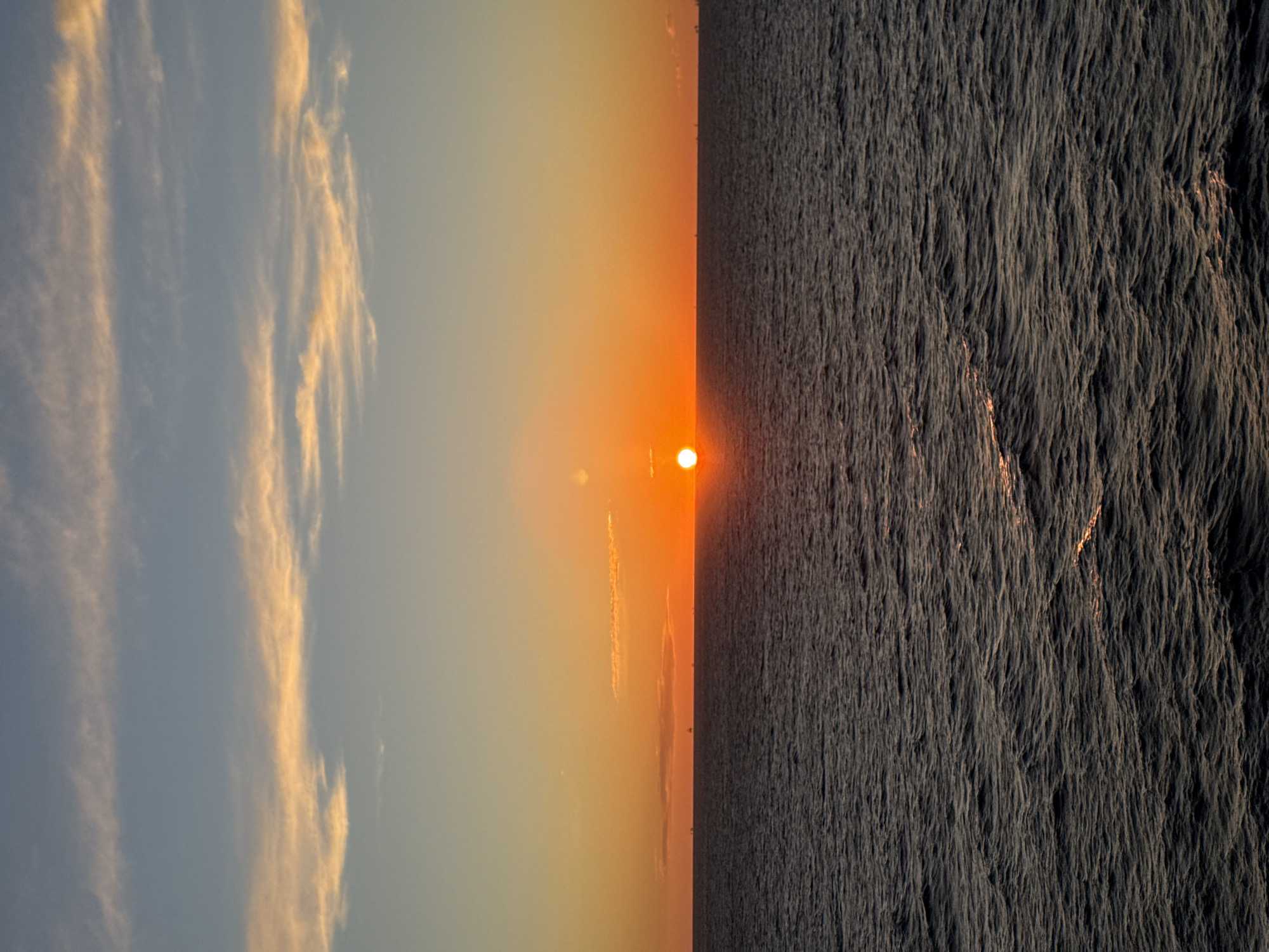A horizontally oriented photograph depicts a sunset scene, likely over a body of water. The left side of the image features a sky with wispy, elongated clouds that are illuminated with a warm, golden hue, transitioning from a lighter blue to a gradient of orange closer to the center. The center of the image is dominated by the setting sun, appearing as a bright, orange orb with a radiating glow. The right side showcases a textured, dark gray expanse, potentially representing the surface of the sea or a similar body of water. The rough texture suggests ripples or waves. No specific location or city is identifiable from the provided image.