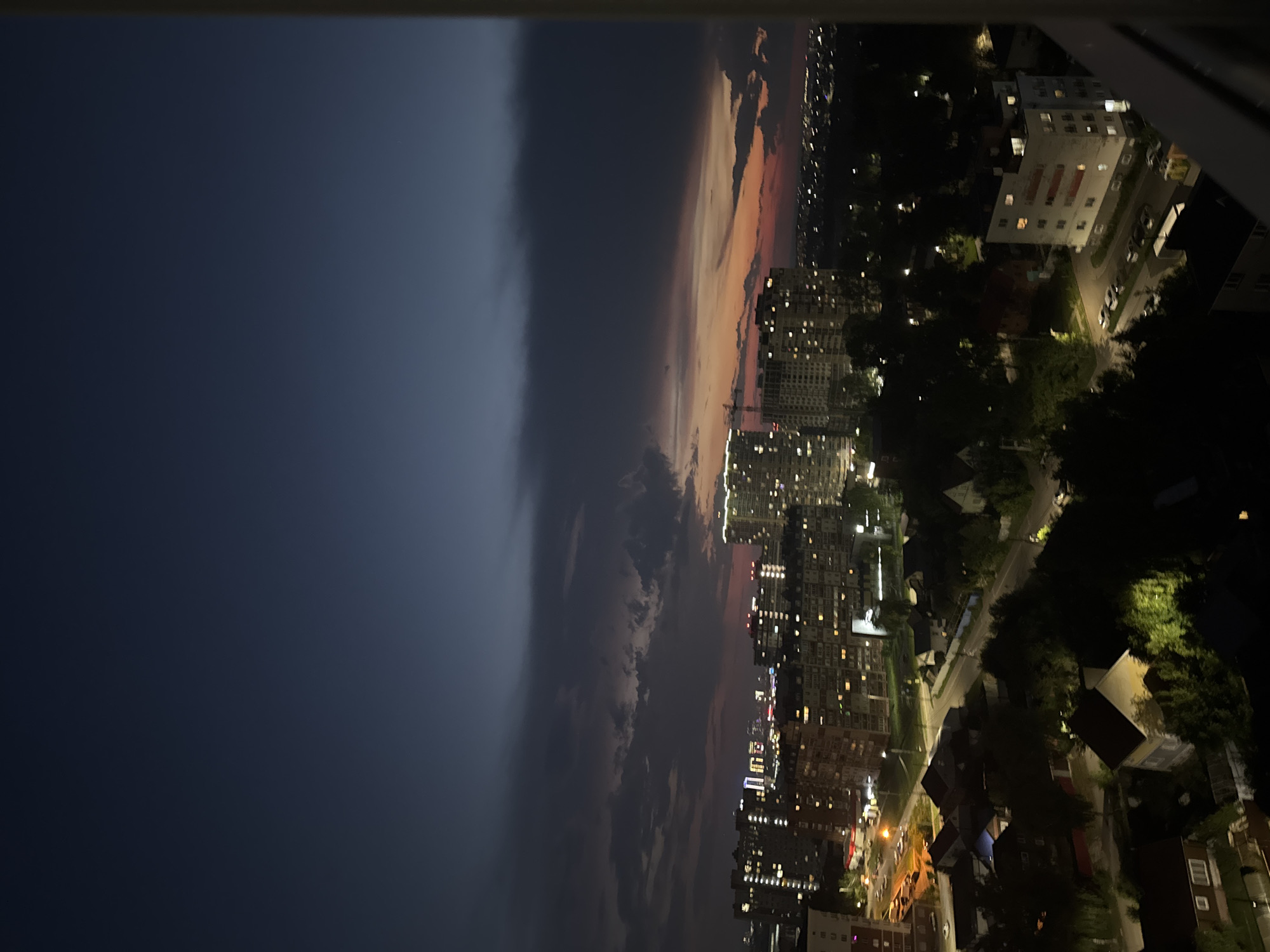 This is a vertical, nighttime, aerial photograph of a residential area in Izhevsk, Russia. The sky is a deep navy blue with dark, streaky clouds in the upper half. The lower half of the sky transitions to a gradient of orange and pink hues, indicative of sunset or twilight. In the middle ground, a cluster of multi-story apartment buildings are illuminated with numerous lit windows, suggesting occupancy. A construction crane is visible between two of these buildings, implying ongoing development. Below the apartment buildings, in the foreground, are smaller houses with pitched roofs, some with visible lights. Streets with cars are present, and trees are scattered throughout the scene, some appearing illuminated by streetlights. The overall impression is a view of an urban residential district at dusk.