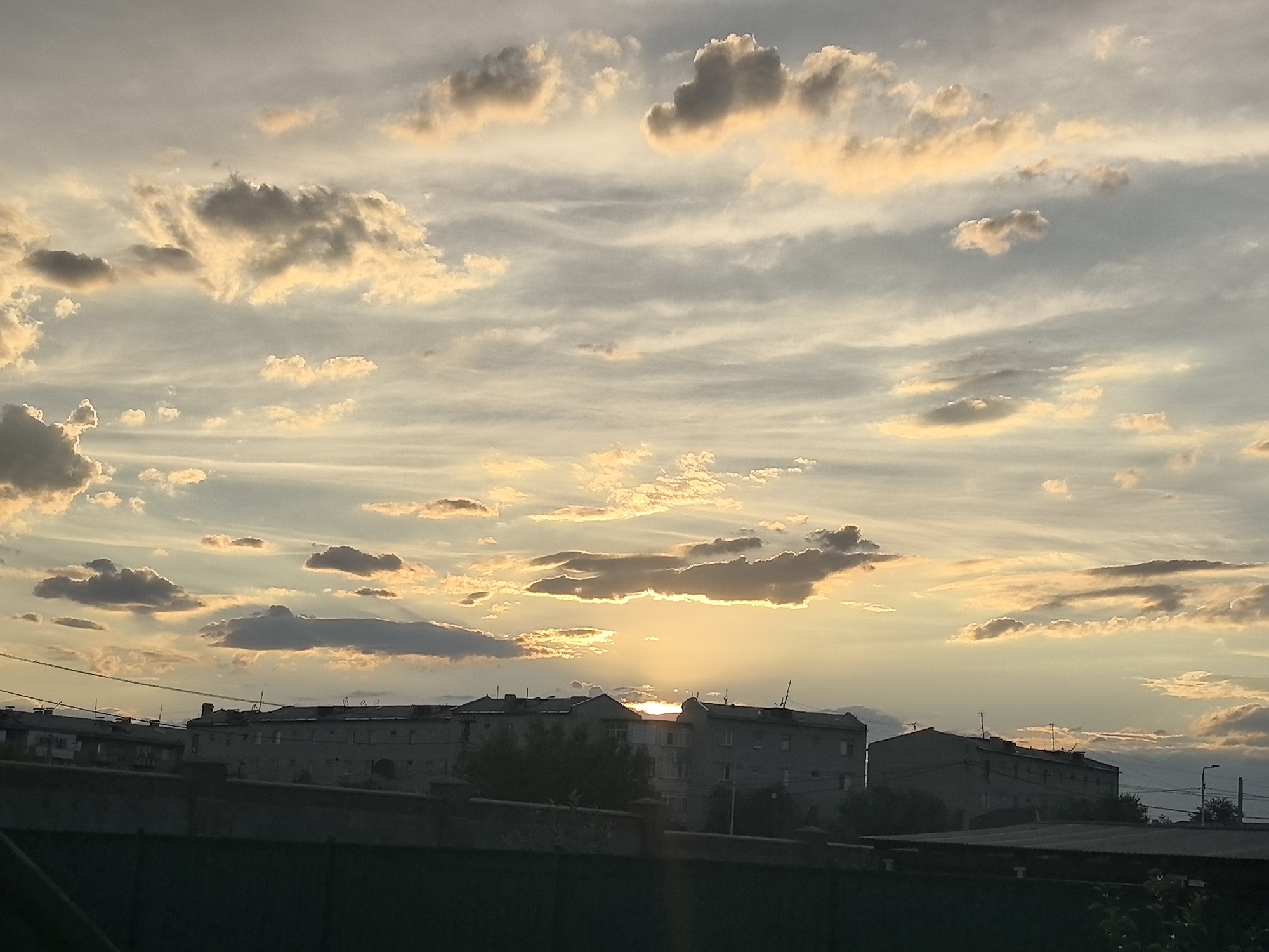 This media file captures a serene sunset scene over the city of Taldykorgan, Kazakhstan. The sky is a canvas of soft hues, with streaks of orange, yellow, and pink painted across a pale blue backdrop. Fluffy, scattered clouds are illuminated by the setting sun, some appearing golden and radiant while others cast gentle shadows. The sun itself is a bright, diffused orb peeking through the clouds, casting a warm glow on the horizon.
Below the sky, the silhouettes of apartment buildings are visible, their flat roofs and repetitive window patterns suggesting a residential urban environment. The architecture appears to be typical of Soviet-era construction, common in many parts of Kazakhstan. In the foreground, a dark, silhouetted fence or wall runs horizontally, obscuring the lower portions of the buildings and adding depth to the scene. Trees are sparsely scattered between the buildings and behind the foreground barrier, their forms softened by the twilight.
The overall atmosphere is peaceful and tranquil, evoking a sense of calm as day transitions into night. The time of day is clearly sunset, with the warm, low-angled light creating long shadows and a dramatic sky. The weather appears clear with scattered clouds, allowing for a beautiful display of natural light. There are no people or specific objects that draw immediate attention, the focus being on the interplay of light, clouds, and the urban landscape. No visible text is present in the image.