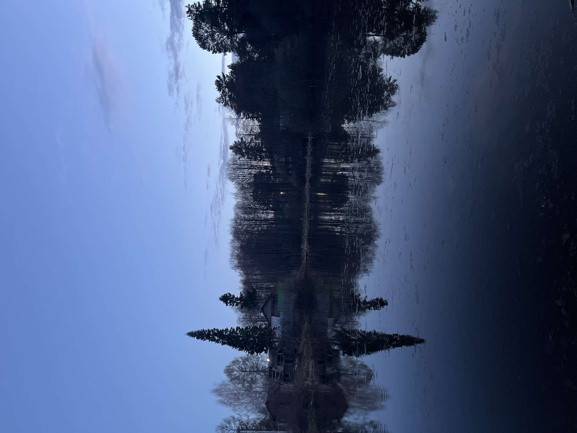 The image depicts a still body of water reflecting a landscape. The sky is visible on the left, transitioning from a light blue hue to darker shades. Scattered clouds are present. The right side of the image shows the reflection on the water, suggesting trees, a building, and other vegetation along the water's edge. Given the prompt's context, this scene is most likely taken in Chaikovsky, Russia.