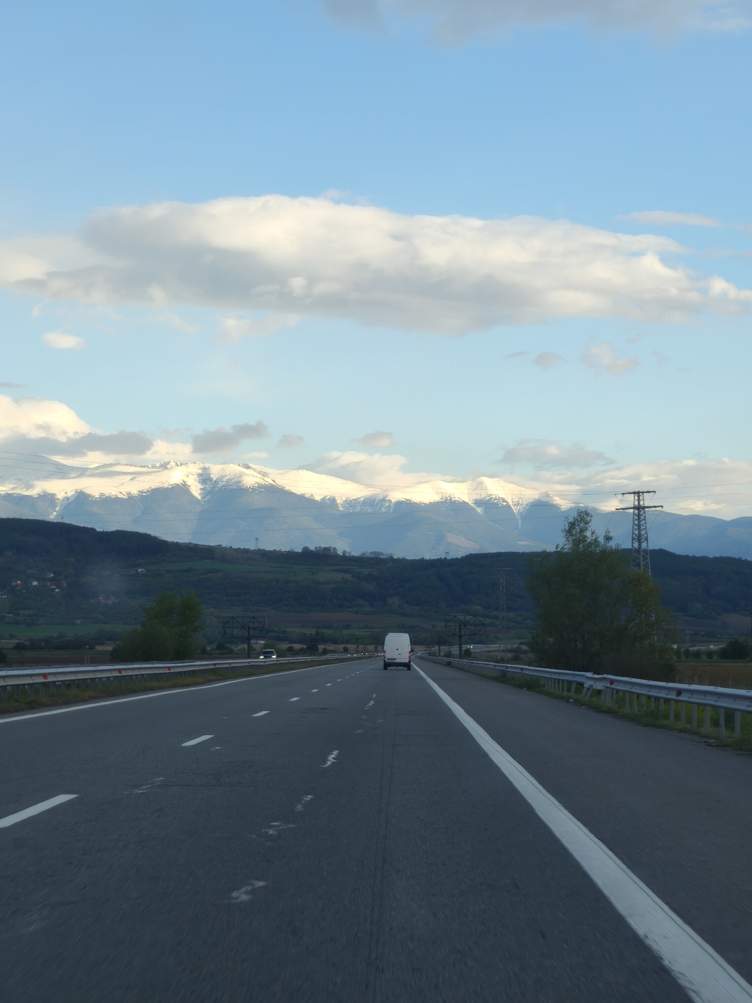 The image shows a multi-lane highway with a white van traveling in the same direction. The road is asphalt and has broken white lines separating the lanes, with a solid white line on the right shoulder. Guardrails are visible on the right side of the road. In the background, there are rolling hills and mountains with snow-capped peaks. The sky is blue with white clouds. A power line is visible on the right side of the highway. The location is likely near Dolna Dikanya, Bulgaria, as indicated.