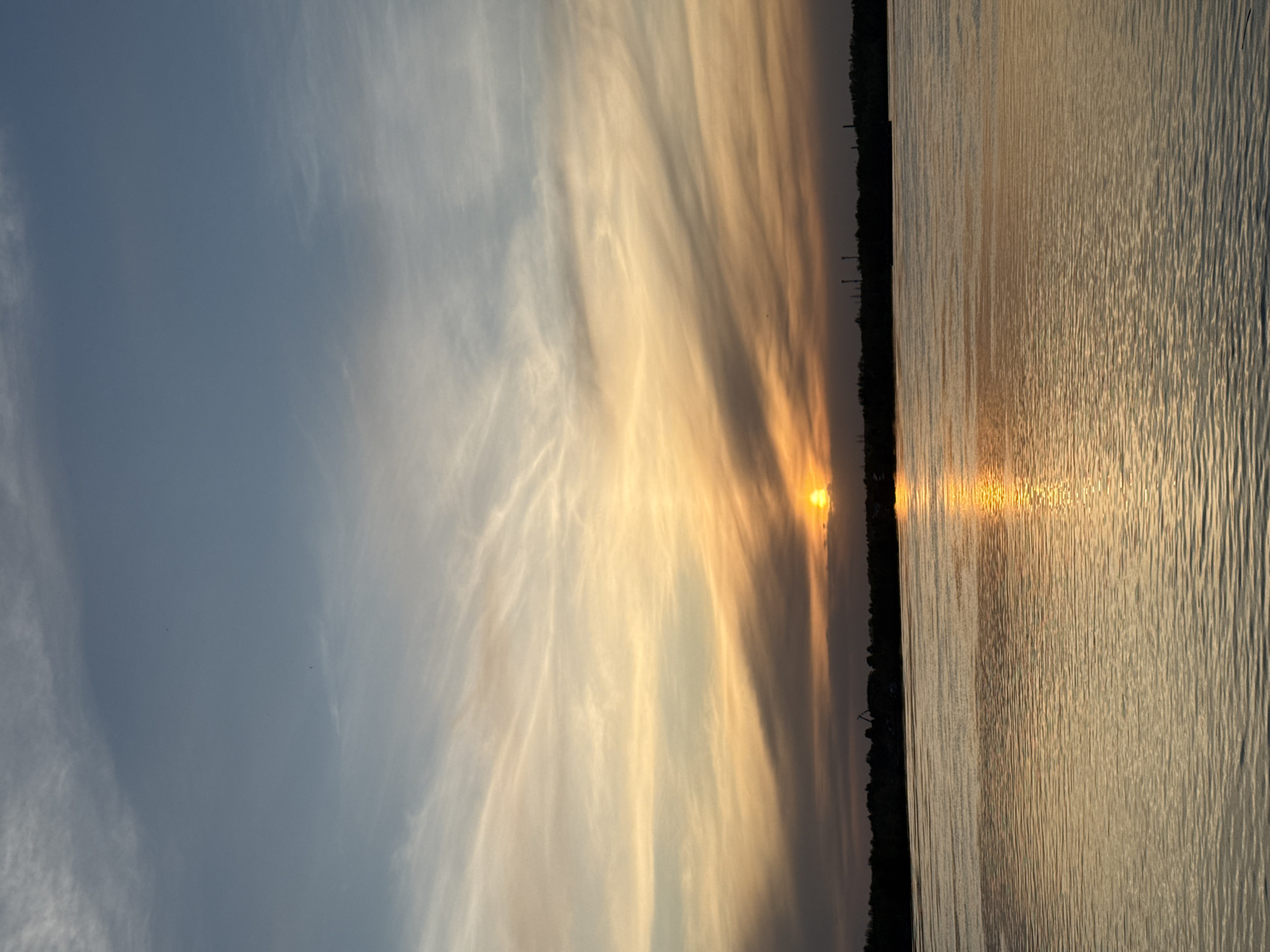 The image depicts a sunset over a body of water, likely the Kama River, in the city of Chaikovsky, Russia. The sun is low on the horizon, reflecting brightly on the water's surface. The sky displays varying cloud formations, transitioning from a clear blue on the left to a yellow-orange near the horizon. A thin line of vegetation and possibly some industrial structures are visible along the shoreline. The overall mood of the image is calm and serene.