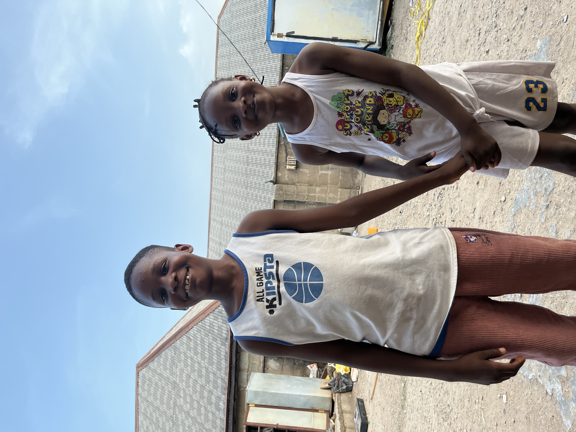 Two young children stand outdoors, holding hands. A girl with braided hair is wearing a white tank top with a colorful cartoon design and white shorts with the number 23 on the side. A boy is wearing a white tank top with a blue basketball logo that says "ALL GAME KIPSTA" and reddish-brown corduroy pants. The background includes corrugated metal buildings and trash. The location is Iddah, Nigeria.