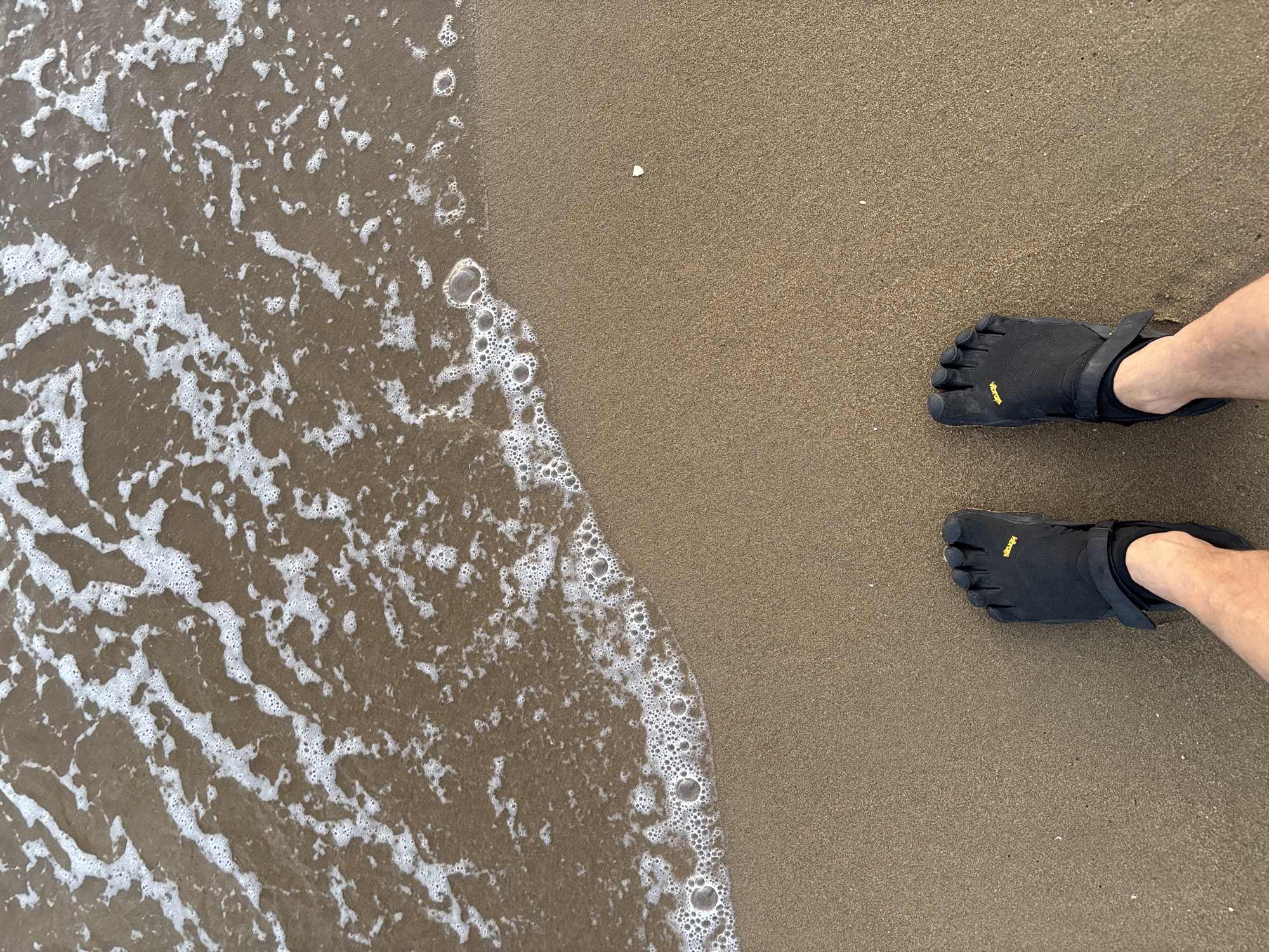 A high-angle, eye-level shot shows a person's feet clad in black Vibram FiveFingers toe shoes standing on a sandy beach in Pattaya City, Thailand. The gentle waves of the ocean are lapping at the shore, creating white foam and small bubbles on the wet sand. The sand is a light brown color, with a few tiny white specks scattered across it. The person's legs are visible from the knees down, with fair skin. The Vibram logo is visible in yellow on the top of each shoe. The scene appears calm and peaceful, likely captured during daylight hours given the lighting.